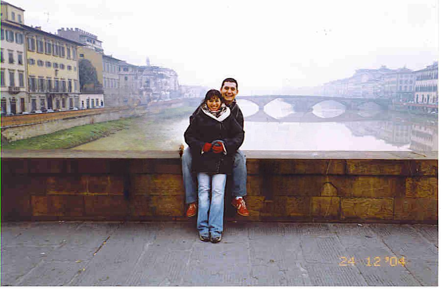 A couple sitting on a stone ledge with a scenic view of an arched bridge and river in the background on a foggy day.