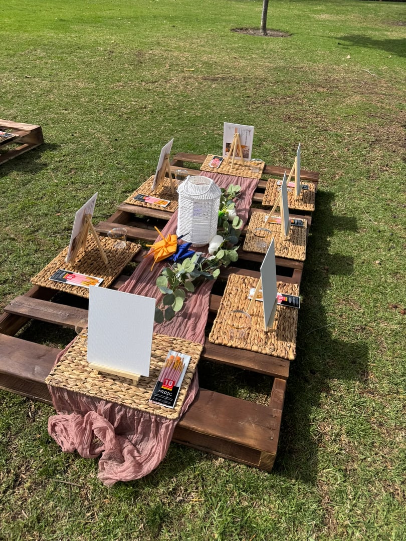 Outdoor art setup with easels, canvases, and art supplies on a low wooden table in a grassy area, decorated with fabric and candles.