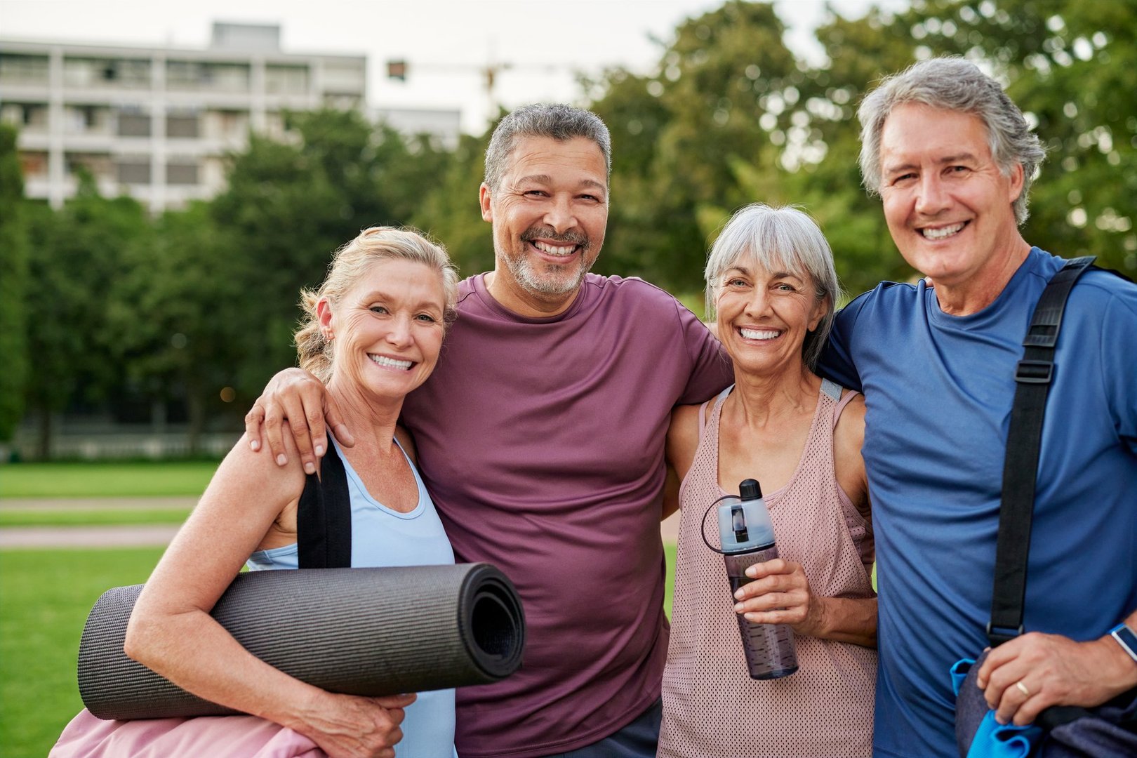 Four seniors smiling and looking at camera after exercising in the park. Multiethnic mature friends enjoying happy moments together with gym bags at park. Group hug and laughter after a fitness session outdoors.