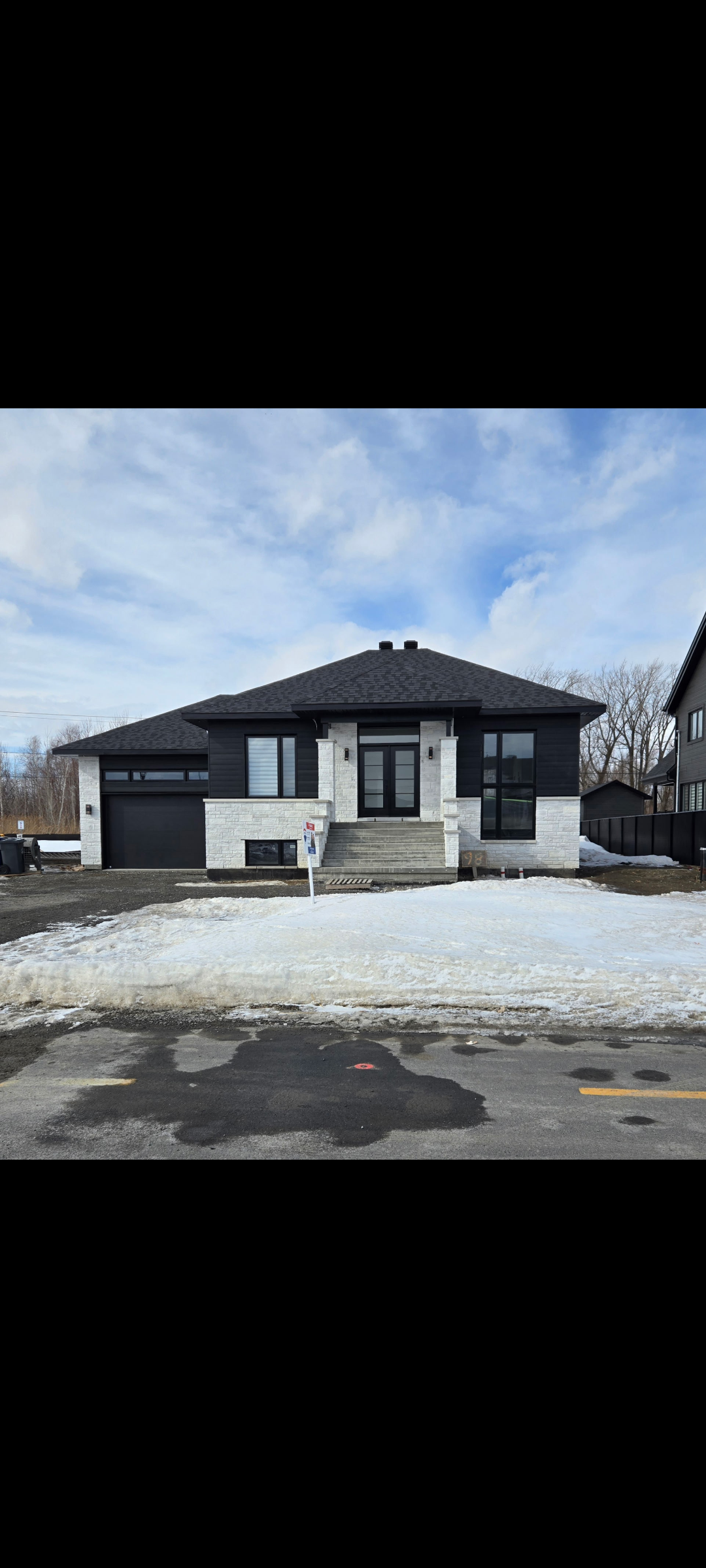 Modern single-story house with a black and white exterior, driveway, and snowy front yard under a partly cloudy sky.