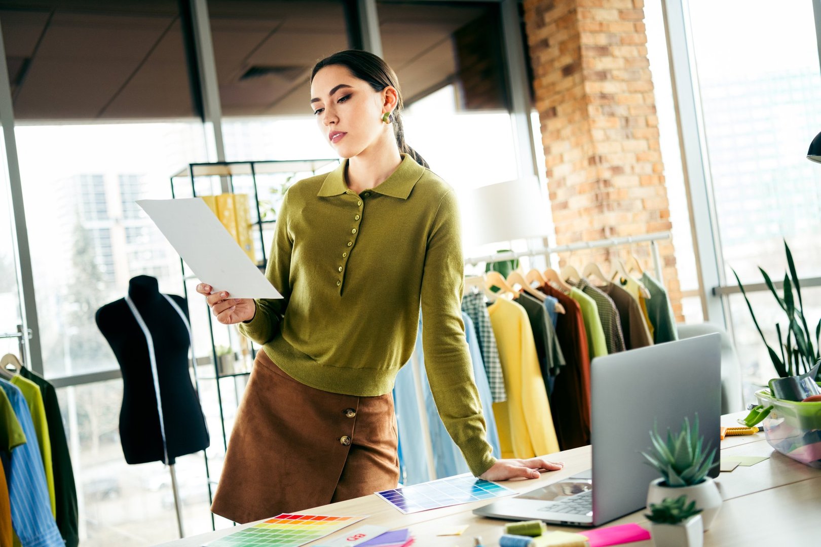 A creative fashion designer works in her atelier, reviewing patterns alongside fabric samples, apparel, and sewing equipment in a bright, stylish workspace.