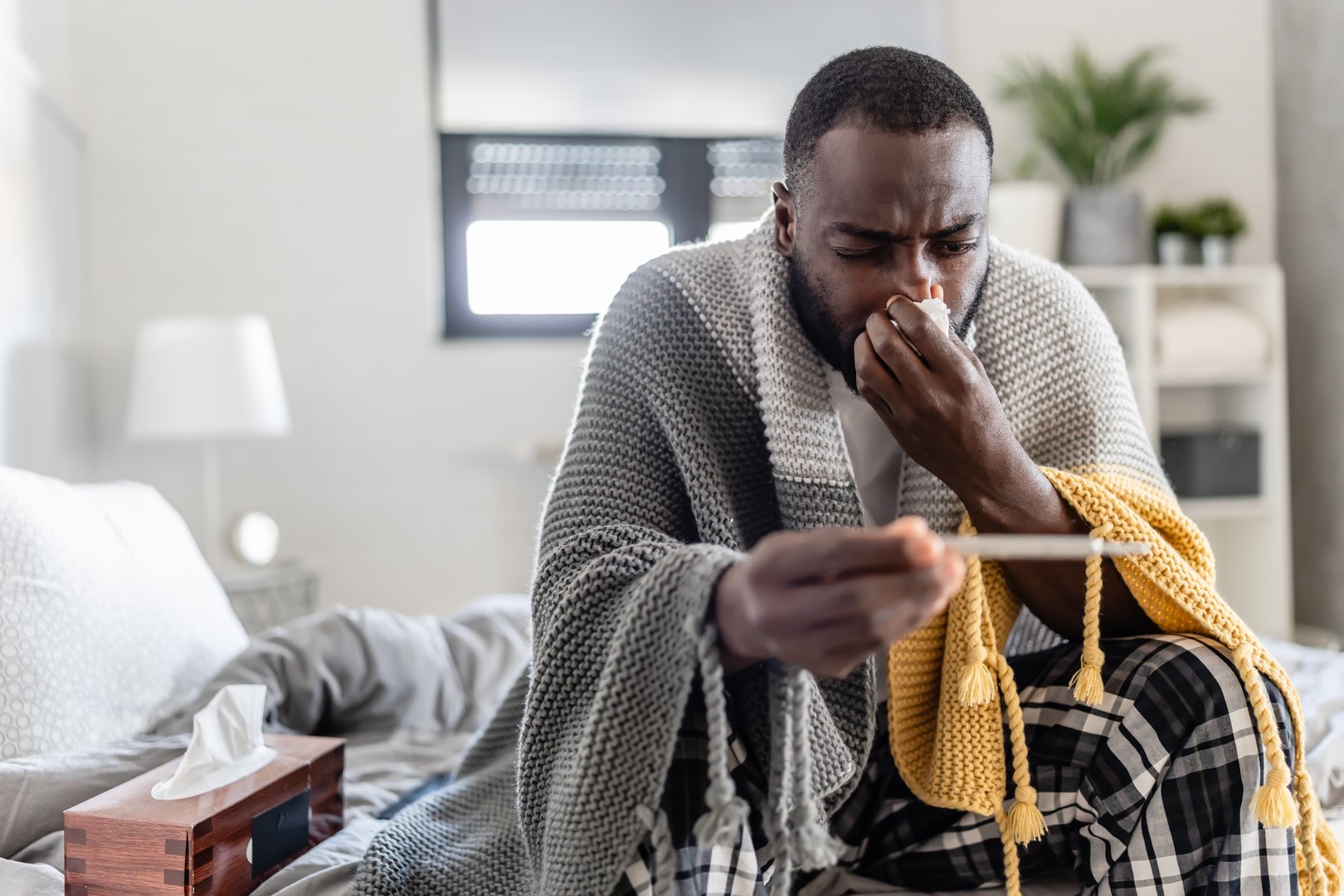 A sick man wrapped in a blanket sits on his bed, coughing into a tissue while checking his temperature with a thermometer. His expression conveys discomfort, illness, and exhaustion.