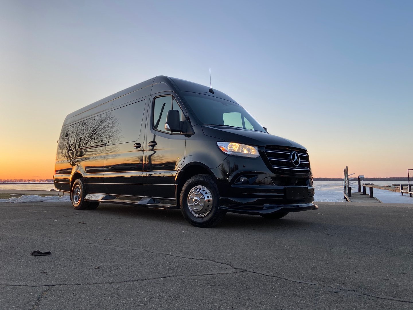 A black Mercedes-Benz Sprinter van parked on a concrete surface with a sunset over a body of water in the background.
