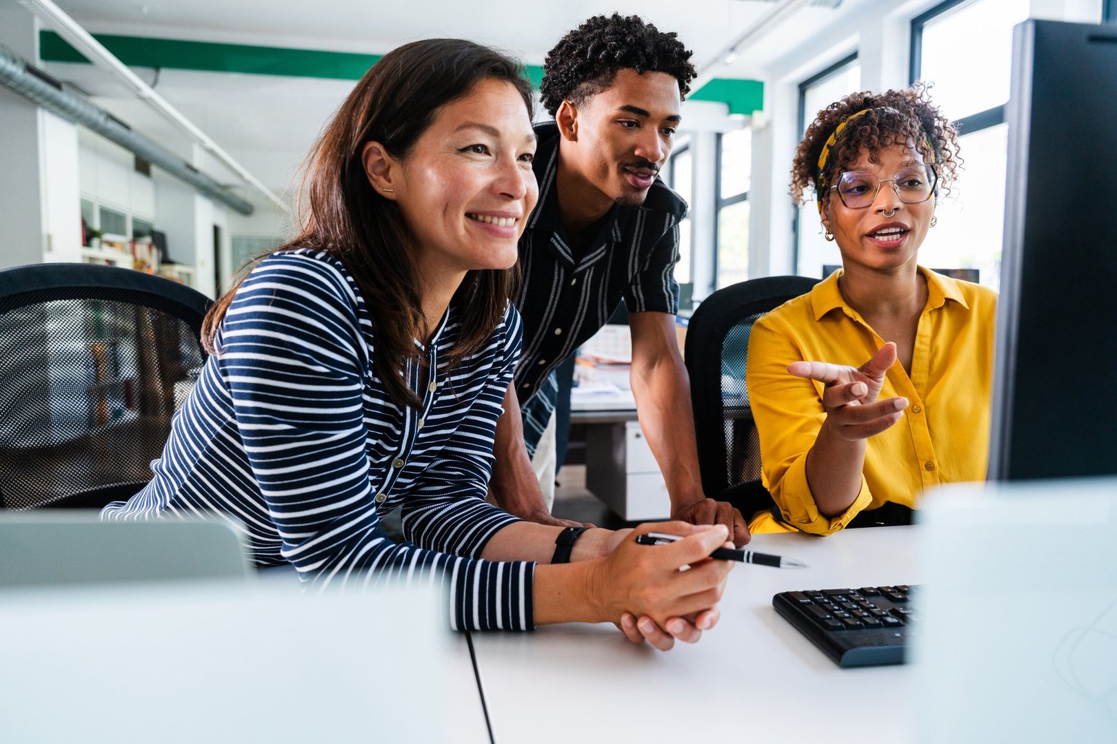 Three smiling business people are collaborating on a project, gathered around a desktop computer in a bright, modern office, discussing strategies and analyzing data