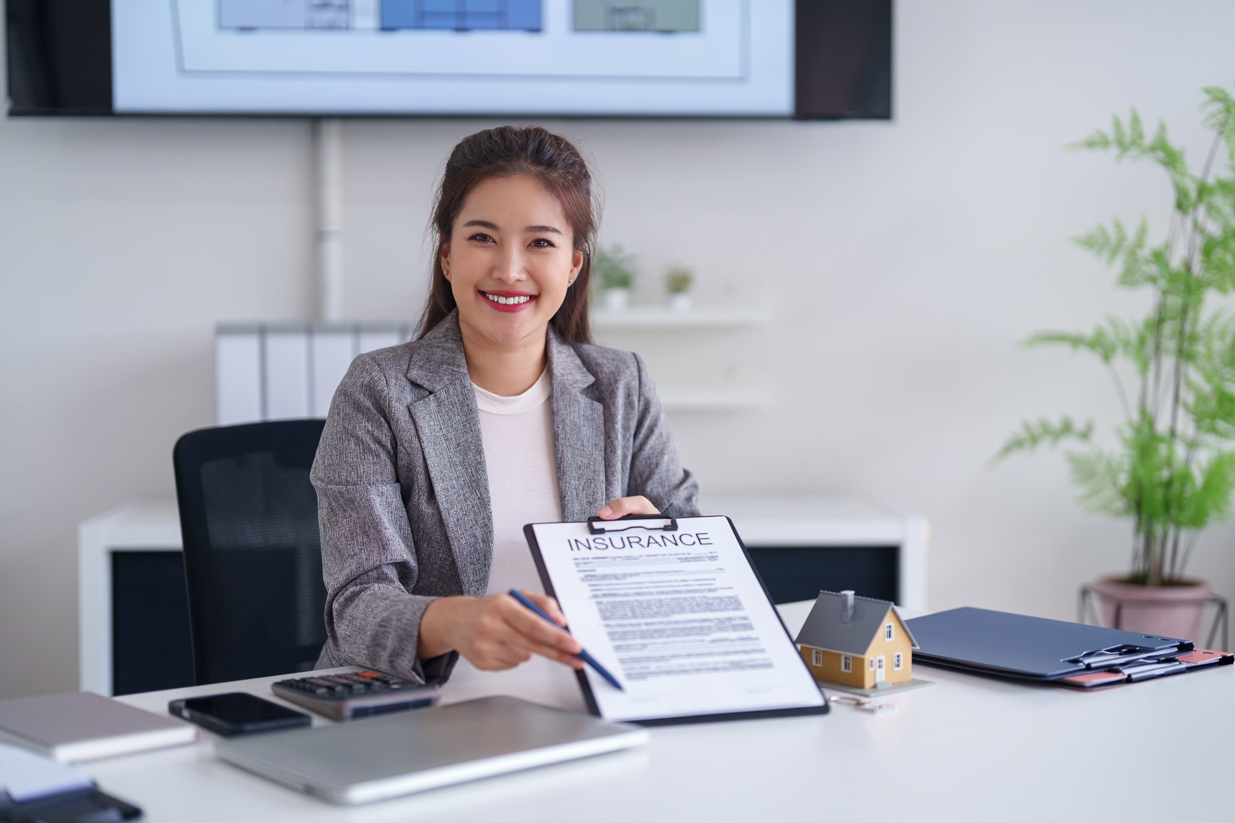 Friendly Asian adult woman insurance agent provides expert advice on home property protection at her modern office desk. Professional service for real estate security.