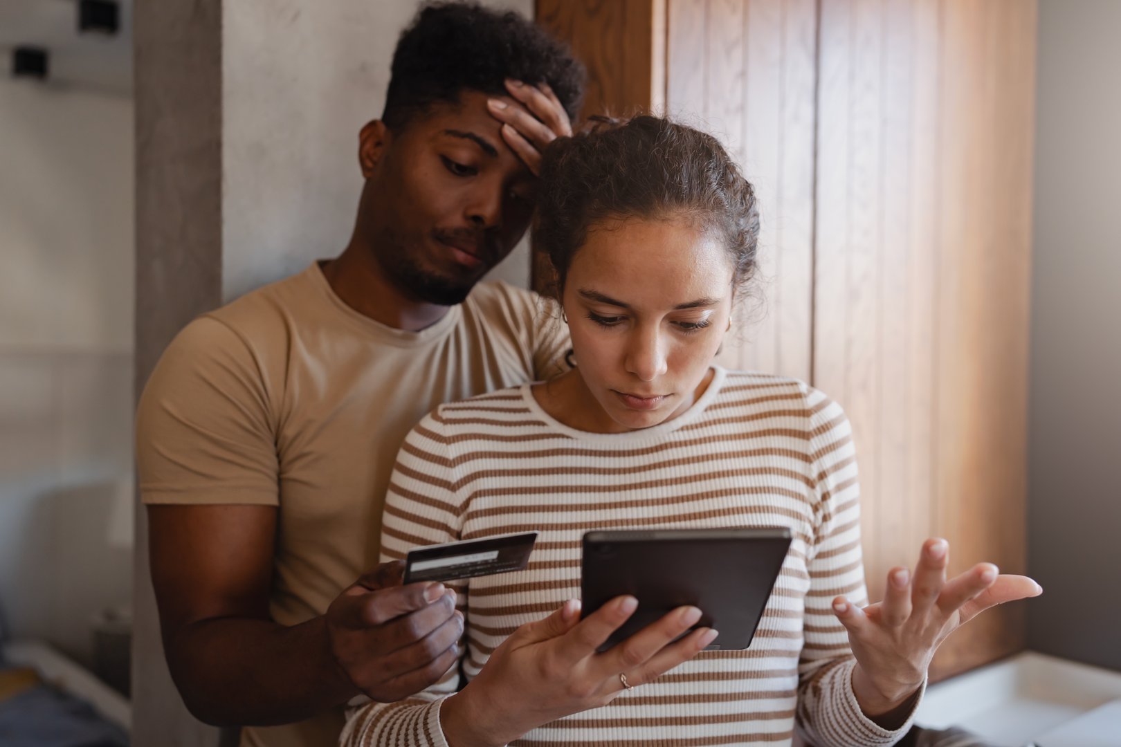 A distressed couple realizes they are victims of online financial fraud, looking worried while checking a tablet and holding a credit card in a modern home setting.