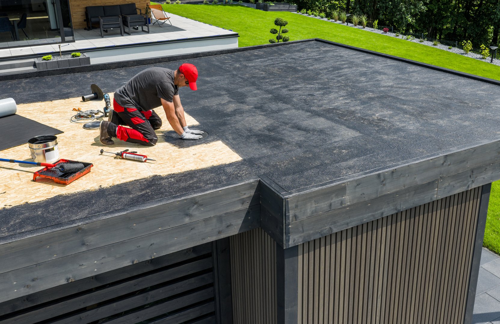 A construction worker applies EPDM roofing material on a flat roof of a modern home in a sunny outdoor environment. Landscaping is visible in the background.