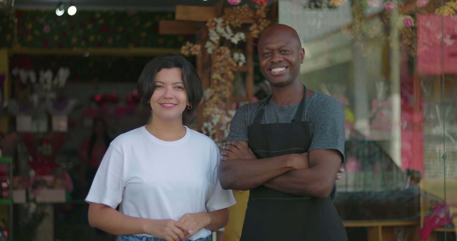 Small business owners standing confidently outside flower shop, smiling man with arms crossed beside woman in white shirt, storefront in background