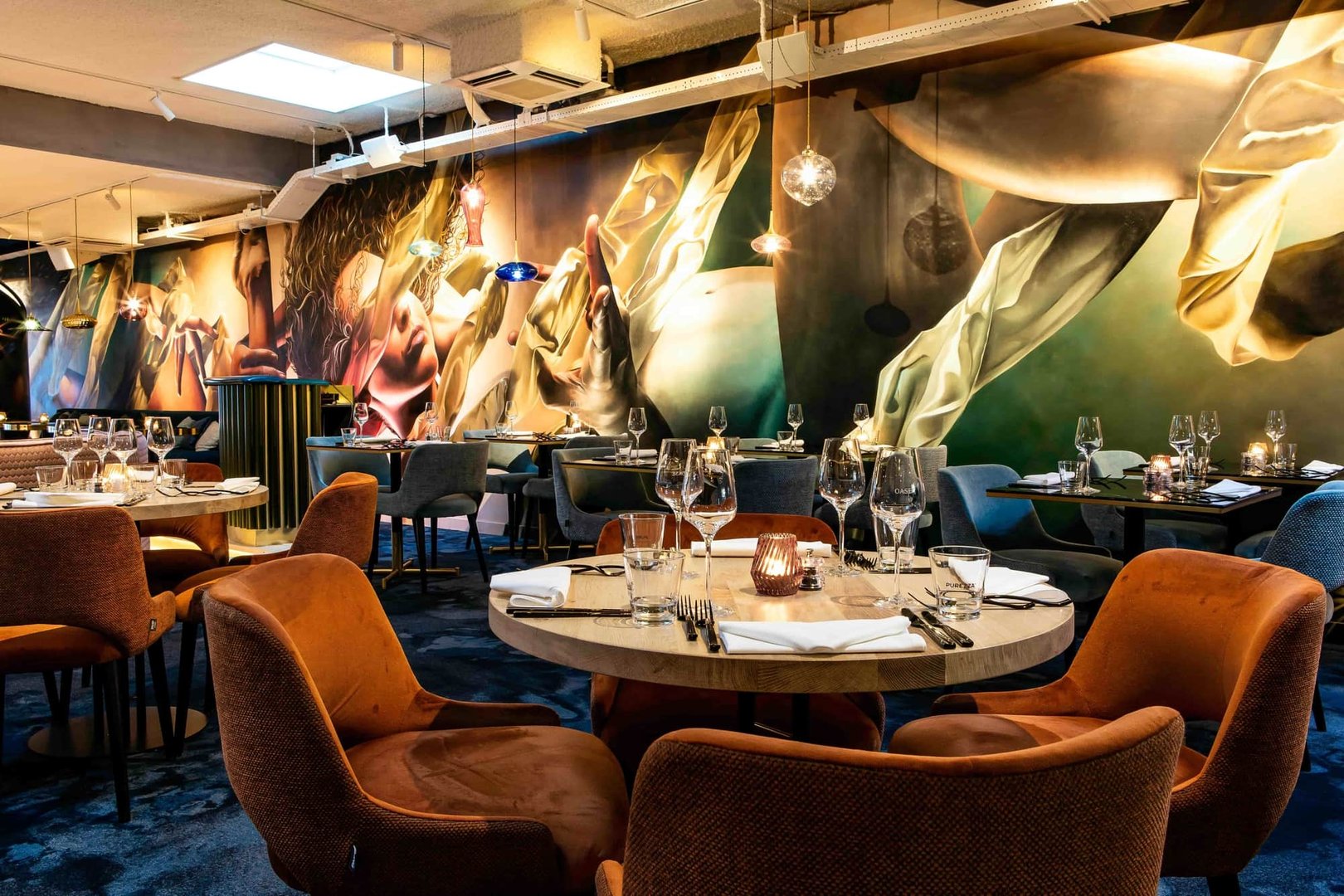 Elegant restaurant with colorful mural, velvet chairs, and tables set with glasses and cutlery.