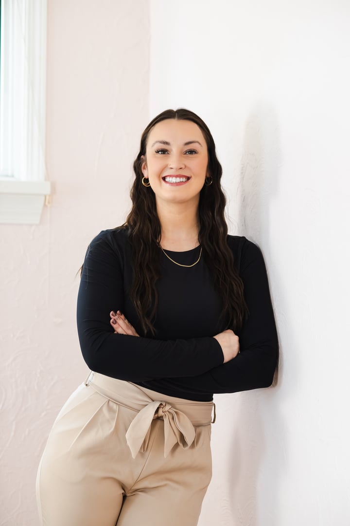 Woman with long hair, in a black top and beige pants, smiling confidently with arms crossed, standing against a white wall.