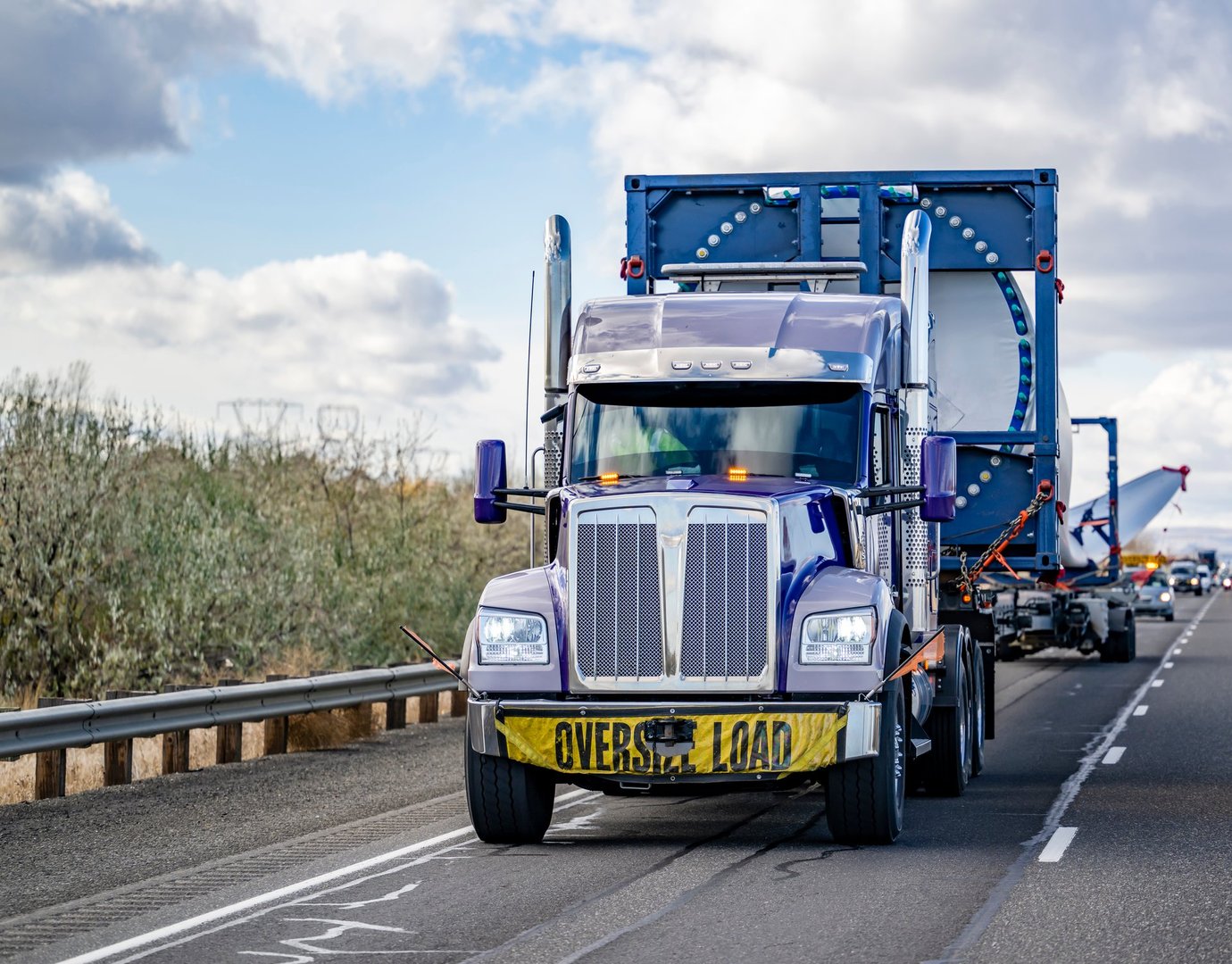 Industrial blue big rig semi truck tractor with additional trolley transports oversize load a super long blade of a wind turbine with an escort car driving on the highway road in Columbia Gorge