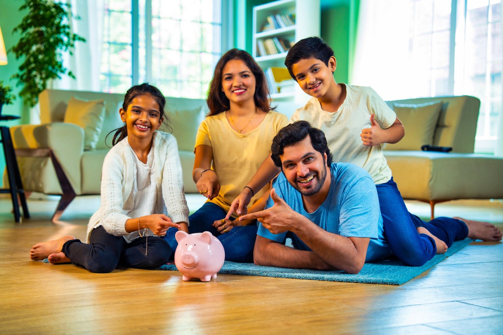 Indian Happy Family of Four in a Modern Living Room Promoting Saving Together, Father Lying on the Floor with Wife and Kids Pointing Toward a Piggy Bank and Smiling