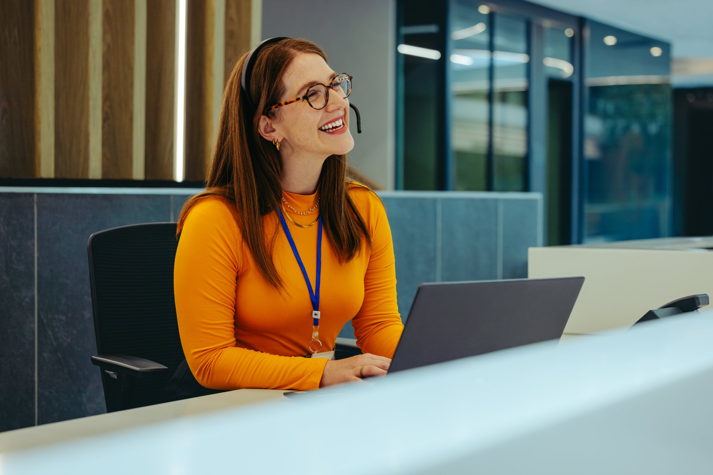 A friendly receptionist in a high-rise office building smiles warmly while assisting clients at the front desk, wearing glasses and a headset, enhancing the welcoming atmosphere.
