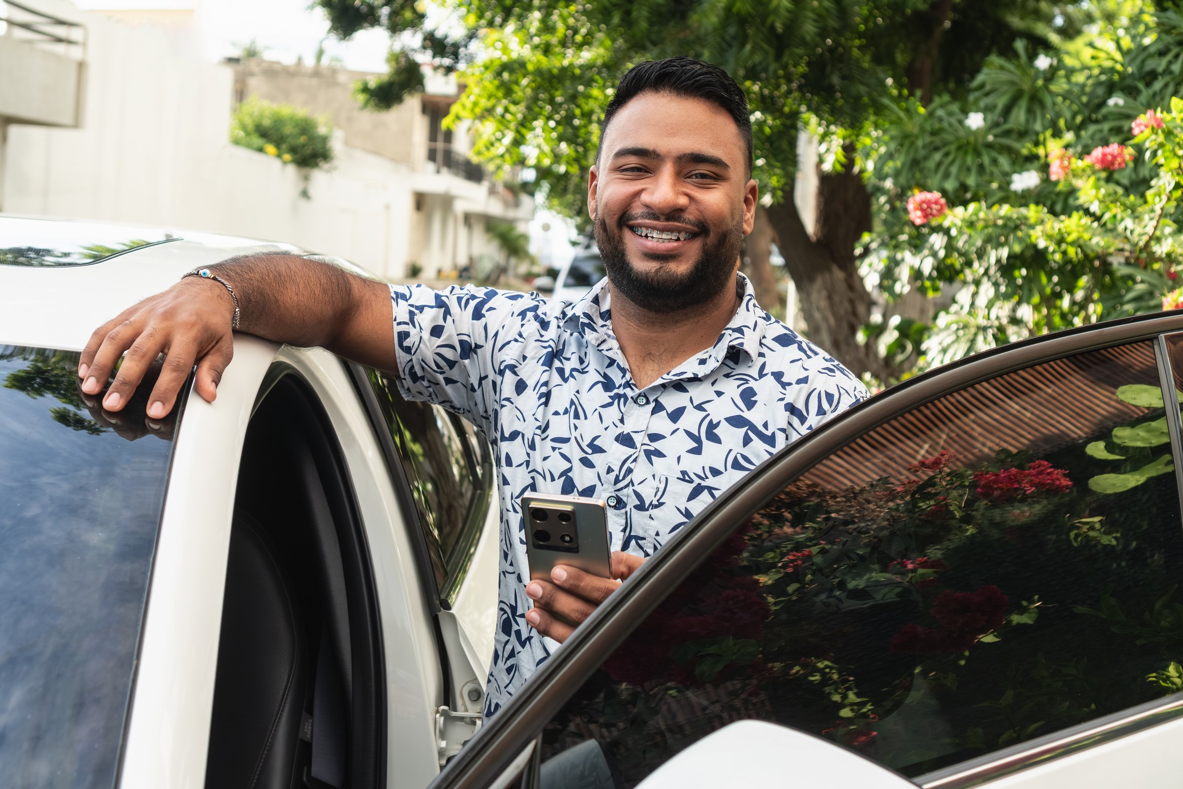 Smiling man standing outdoors by open car door holding cell phone and looking at camera