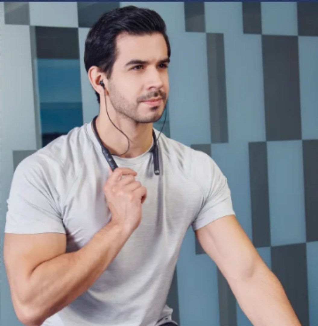 Man in a gray T-shirt wearing earphones, sitting indoors against a geometric patterned wall.