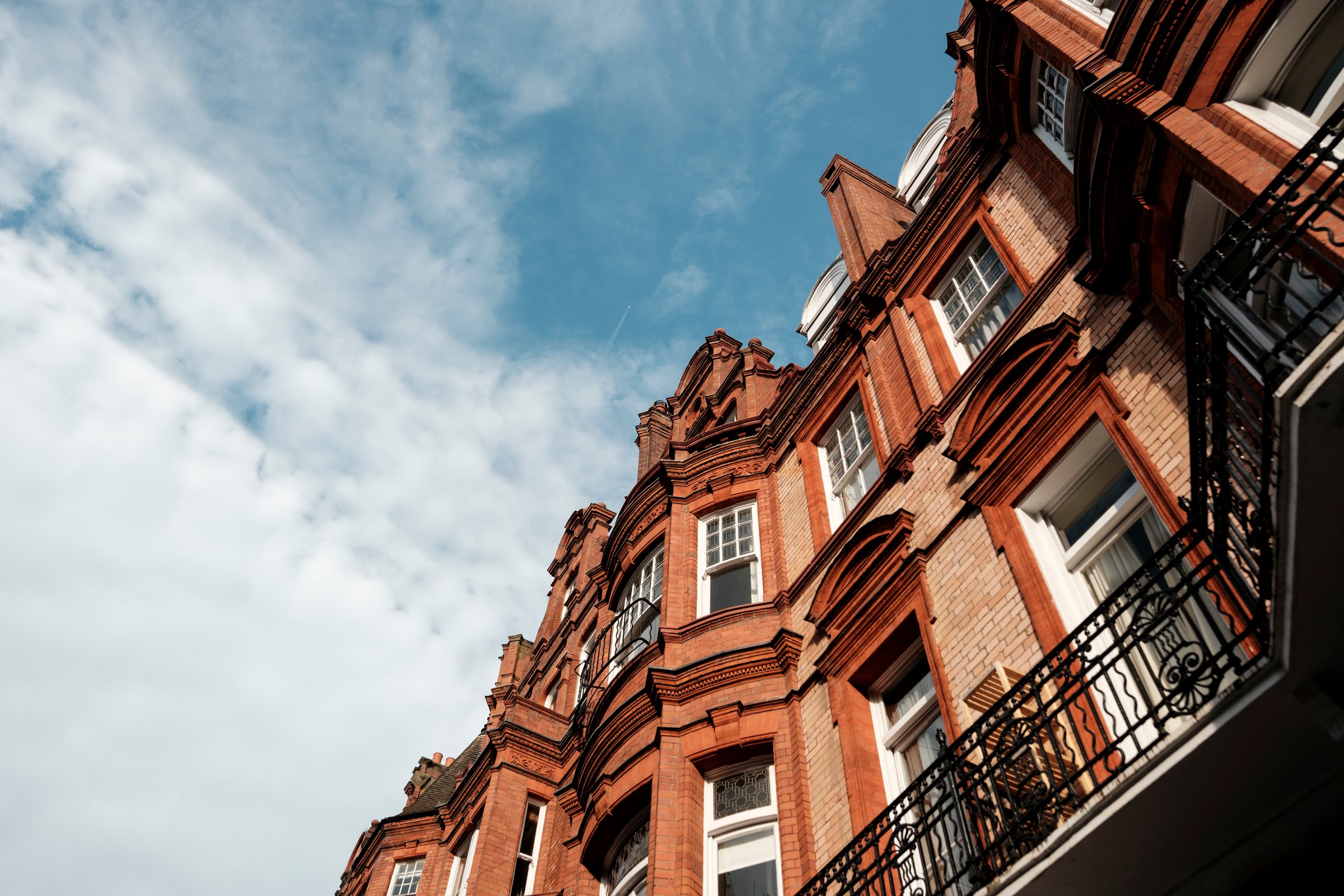 Ornate red brick victorian buildings featuring bay windows and balconies, displaying classic london architecture under a bright blue sky with wispy clouds