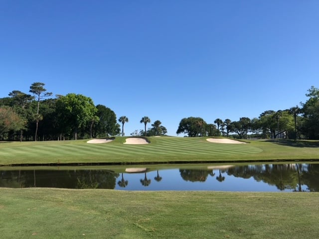 Golf course with green fairway, sand bunkers, and a reflecting pond under a clear blue sky.