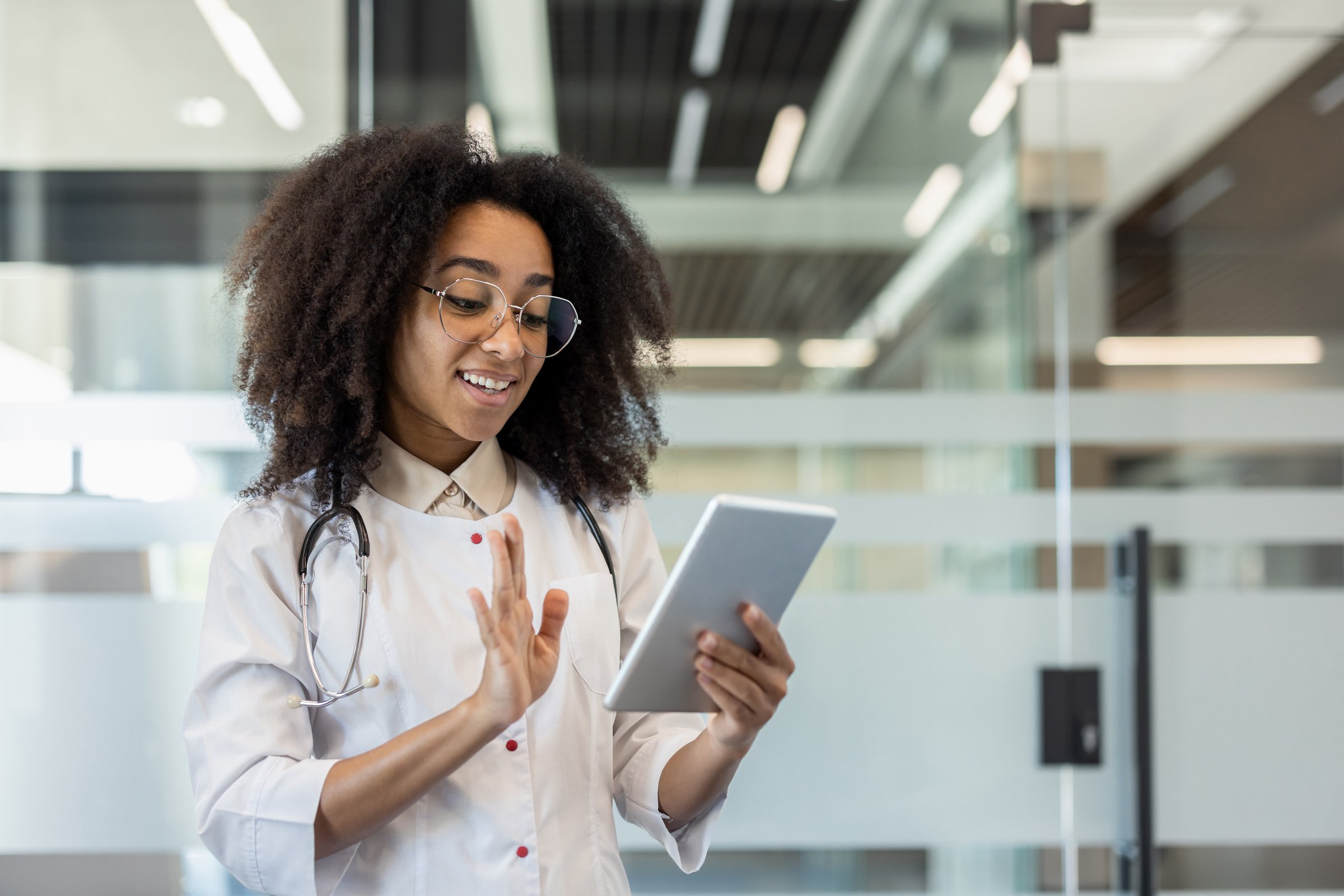 A young African-American female doctor stands in the clinic in a white coat and talks on a video call to the camera of the tablet she is holding in her hands.