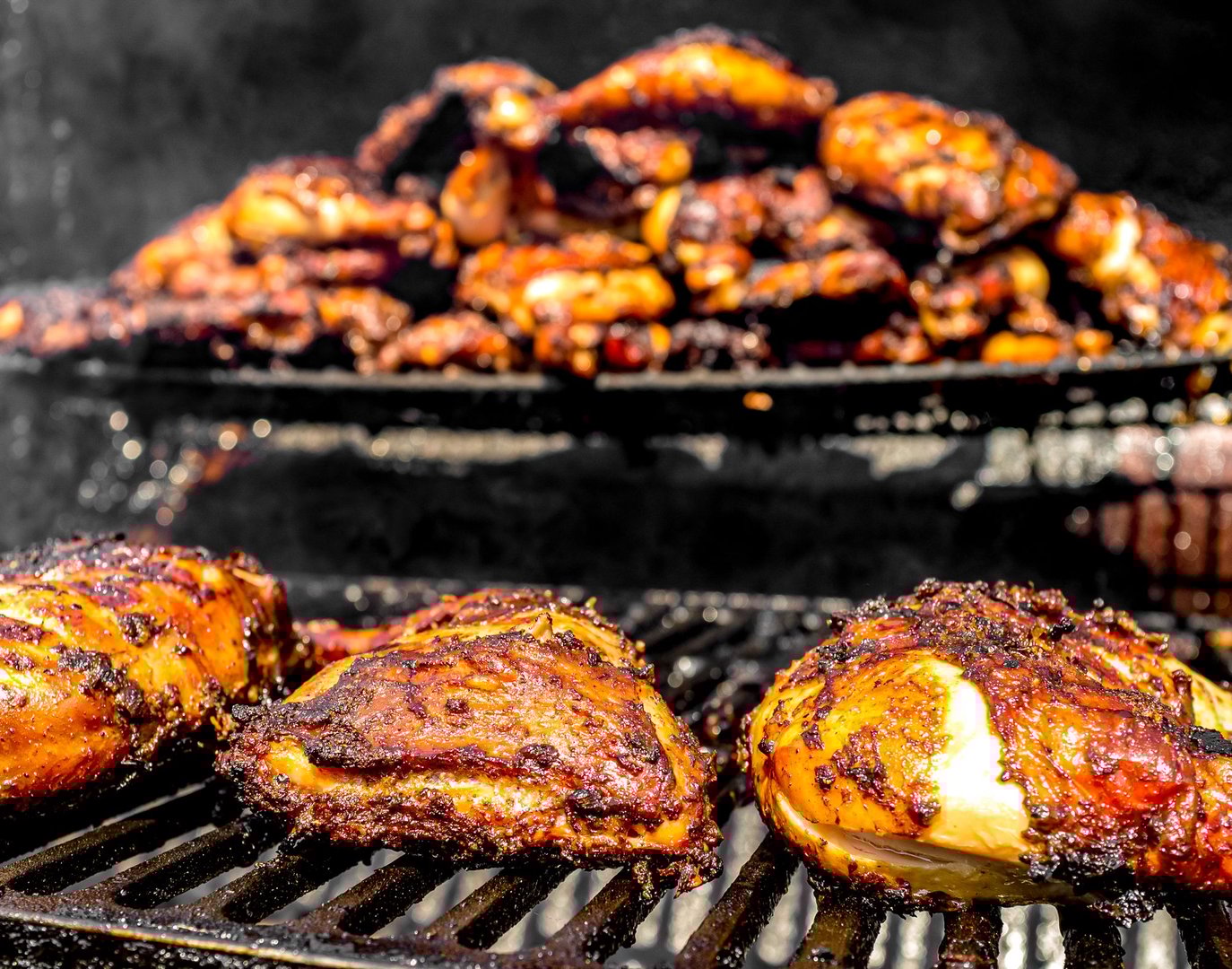 Grilled chicken pieces with crispy skin cooking on a barbecue grill, with more chicken in the background.