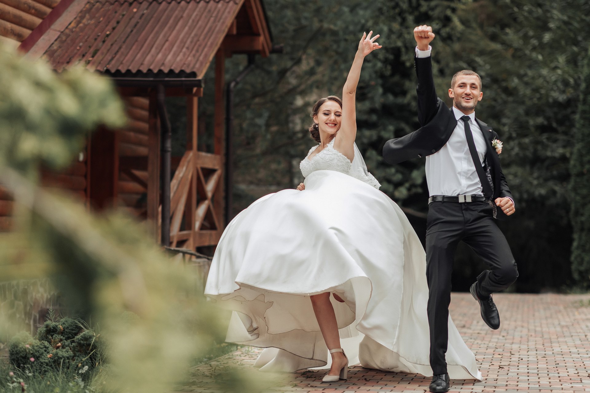 A bride and groom are jumping in the air, celebrating their wedding. The bride is wearing a white dress and the groom is wearing a black suit. The scene is set in a forest