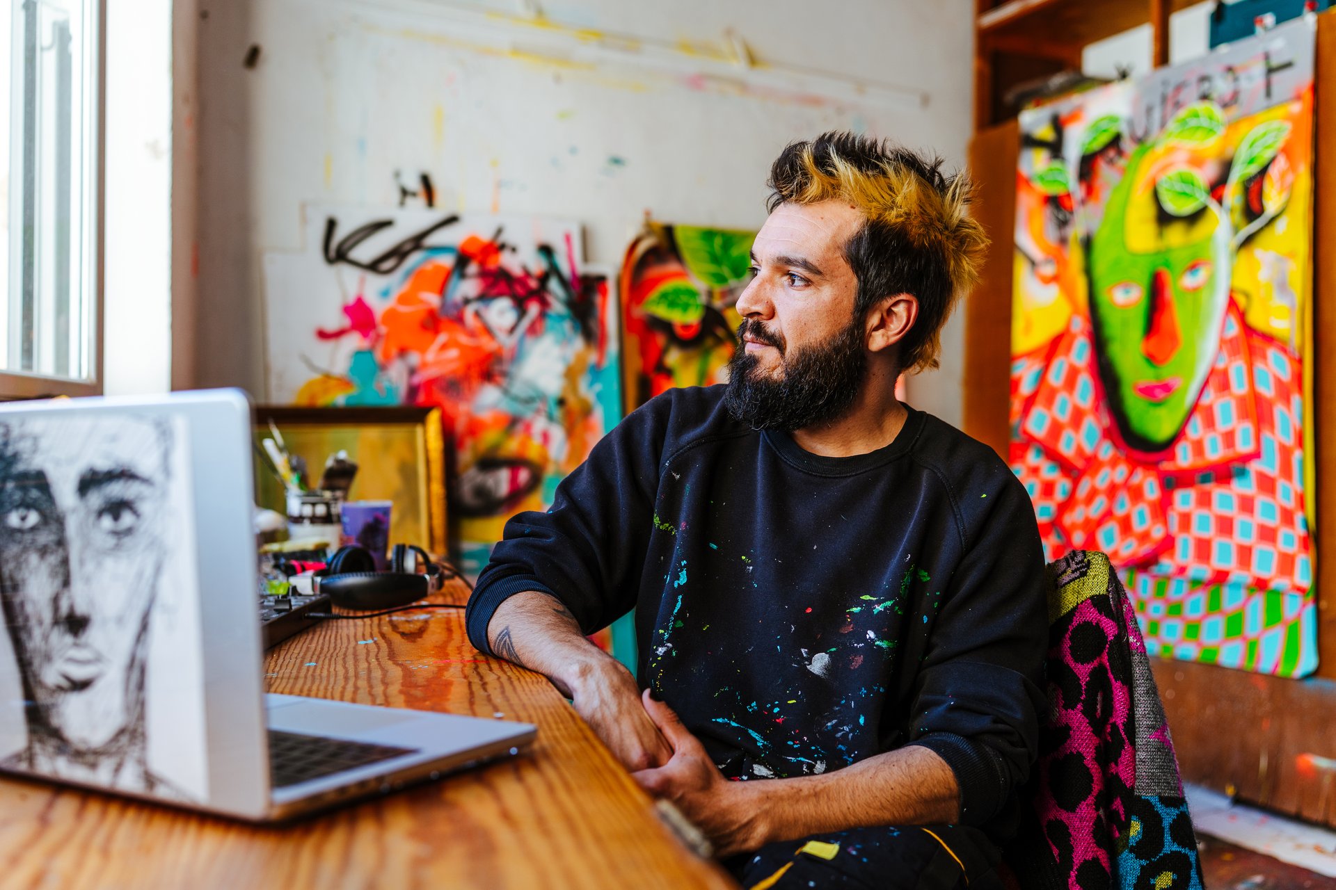 An Argentine artist sits at his workspace, gazing thoughtfully while surrounded by abstract paintings and creative tools in his vibrant studio