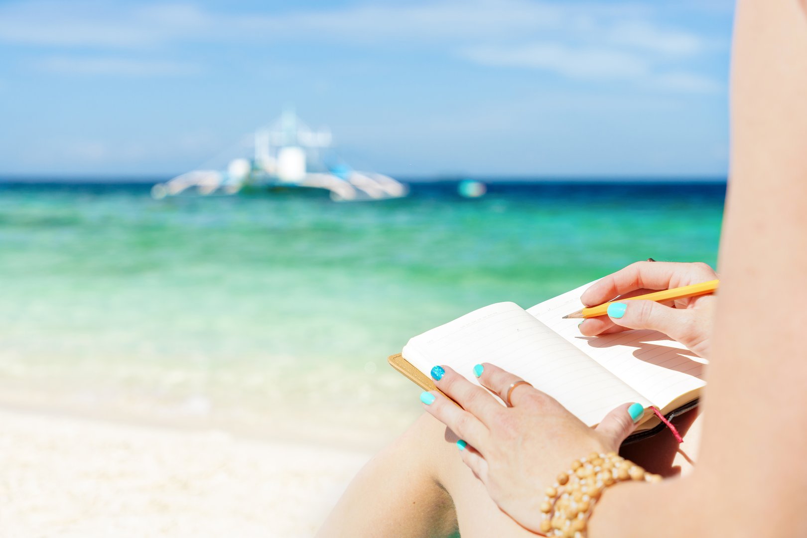 european woman is sitting on the coast of tropical turquoise sea and wrigting by pen in notepad at sunny summer day under blue sky
