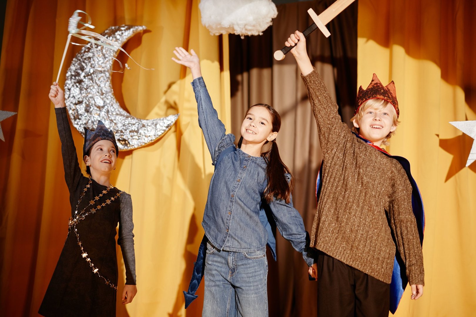 Group of children performing during holiday celebration on stage decorated with giant stars, wearing festive costumes and holding props, showing enthusiasm and joy