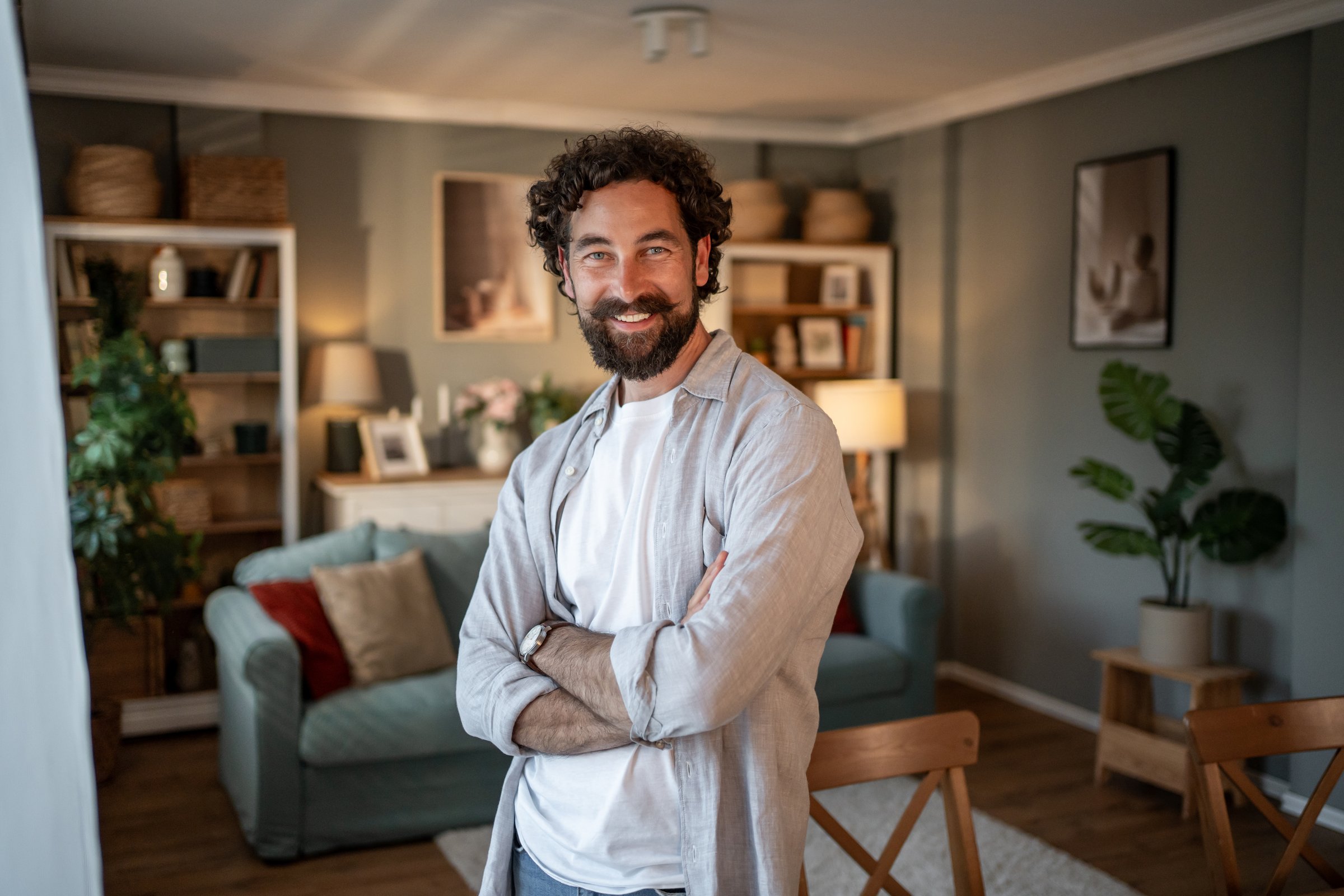 Mid adult man with curly hair and a beard is standing with his arms crossed, smiling at the camera in his modern, stylish apartment living room