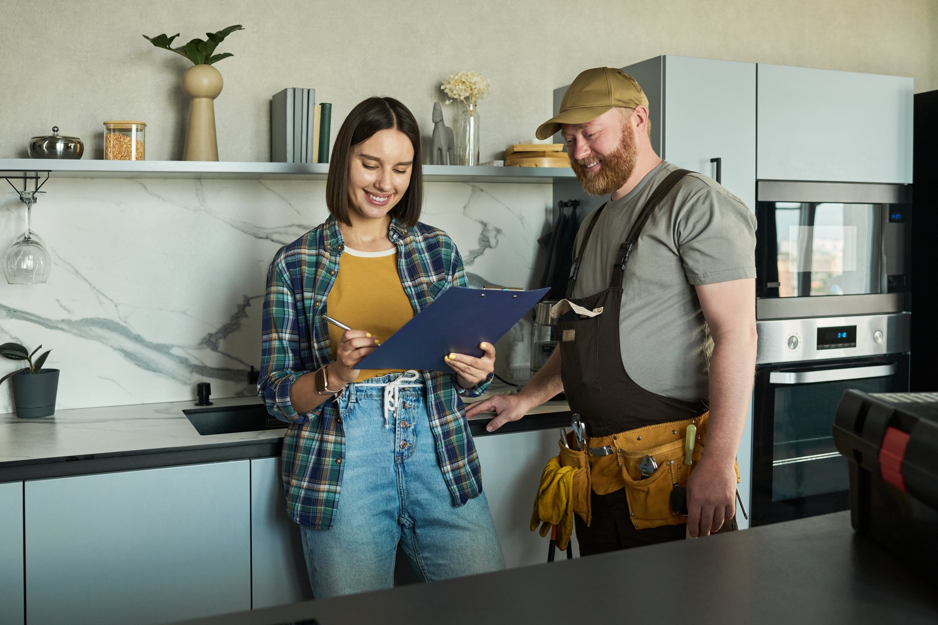 Caucasian young adult woman reviewing documents with Caucasian middle aged man wearing work uniform in modern kitchen, both standing and smiling while discussing project details