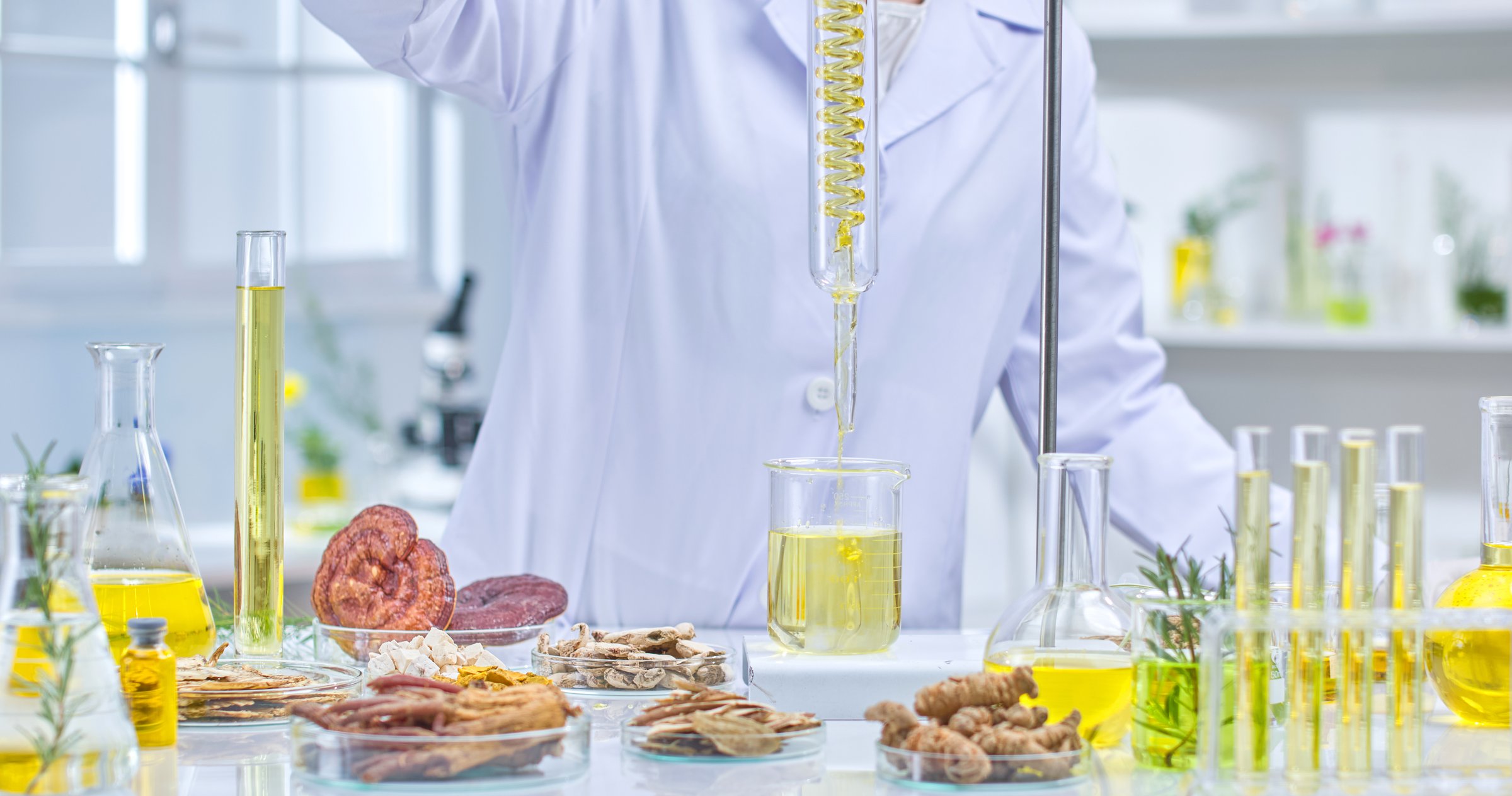 A scientist wearing a lab coat, gloves and a protective mask is holding a flask filled with a yellow liquid, examining it in a lab surrounded by herbs, flasks and other lab equipment.