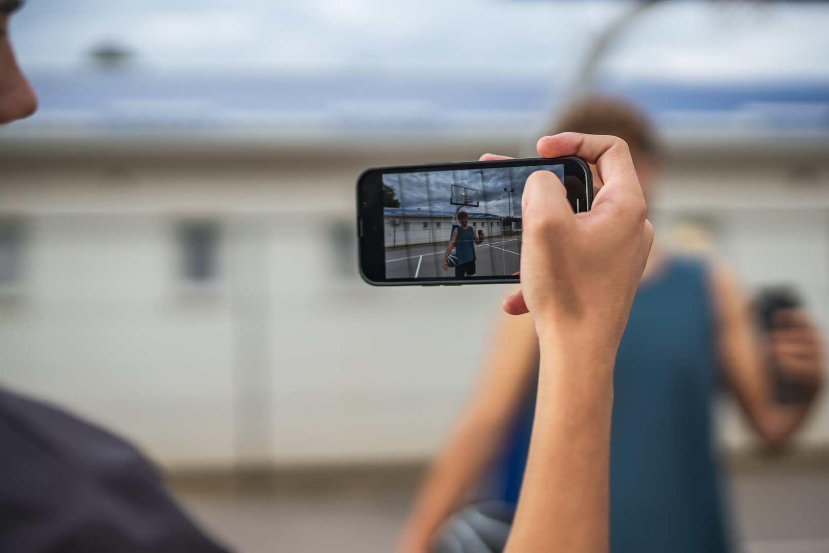 Person holding smart phone filming a young man posing with a basketball on an outdoor urban court under a cloudy sky, capturing casual sports lifestyle content for social media