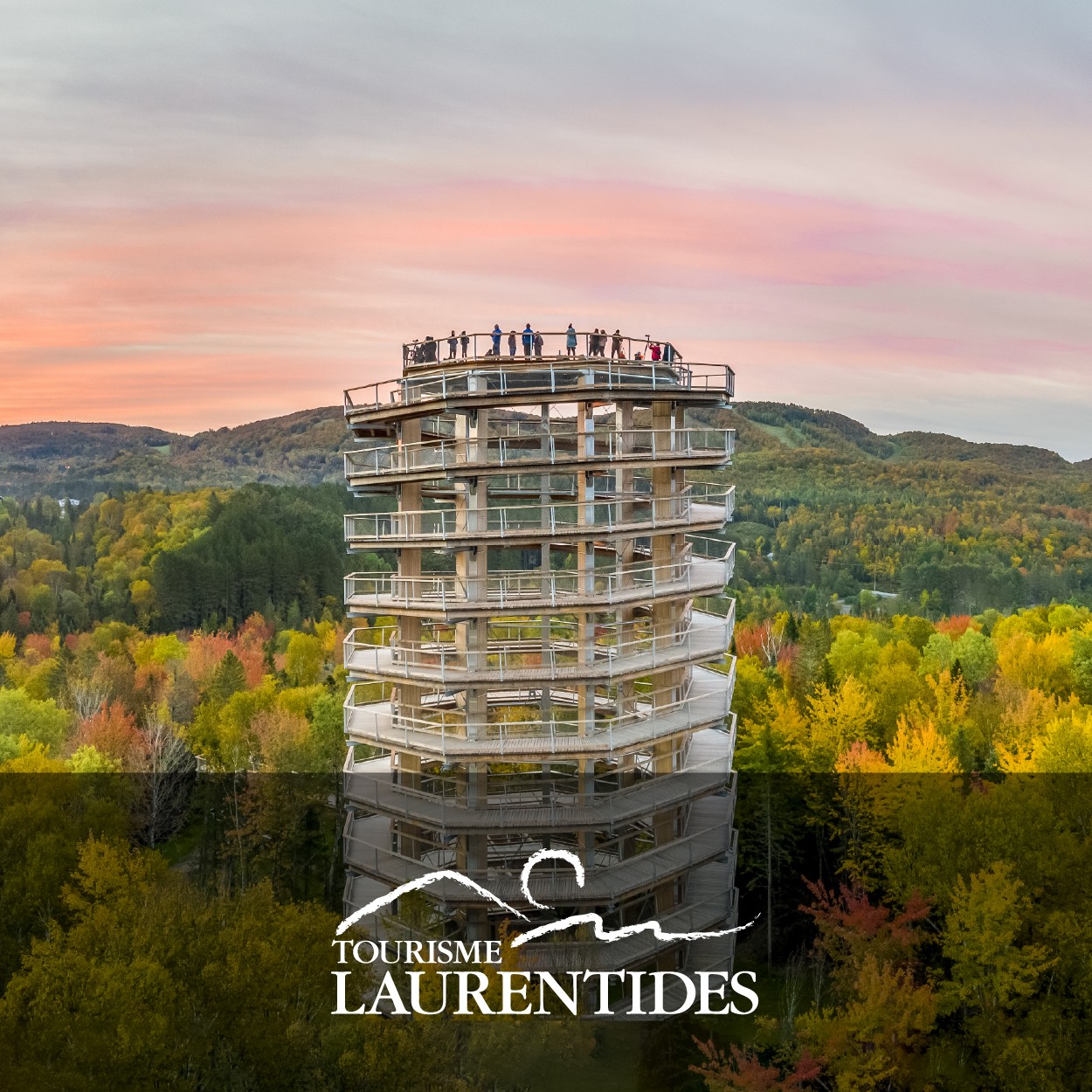 Tower with spiral walkway surrounded by autumn trees and hills, people at the top, under a colorful sunset sky.