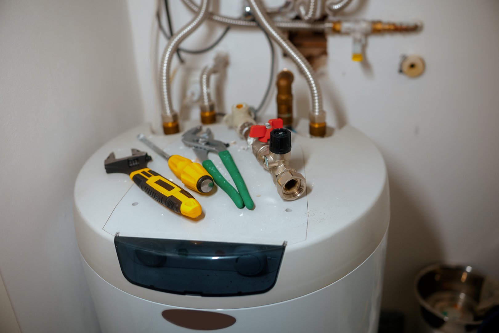 An adjustable wrench, screwdriver, and pliers rest on a white water heater, surrounded by pipes and valves, signaling active plumbing repair or maintenance work on a domestic heating system.