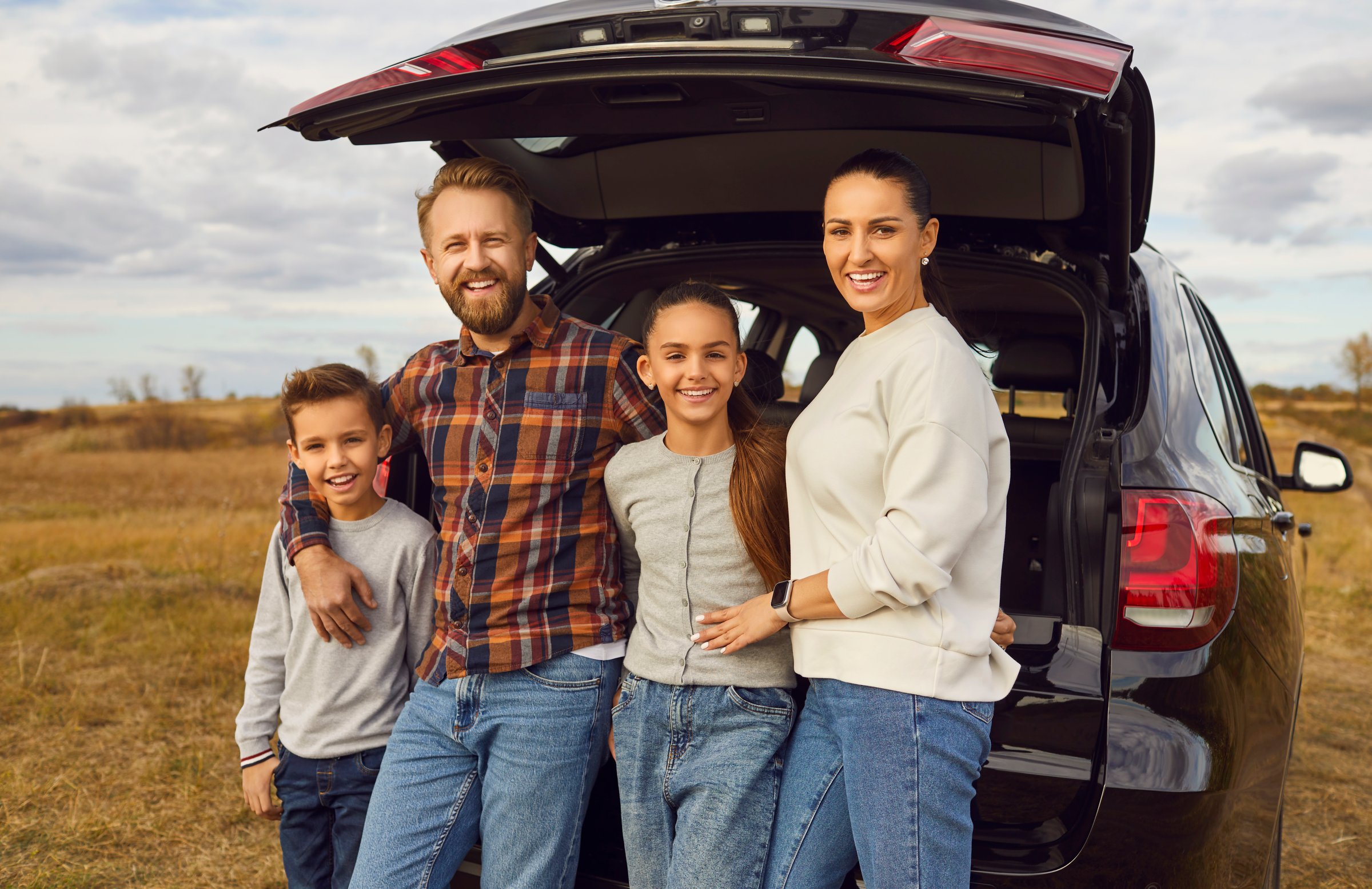 Family with modern automobile enjoying road trip adventure together. Happy loving mum, dad and children standing by open car trunk during roadtrip in rural fields on cloudy autumn day. Travel concept