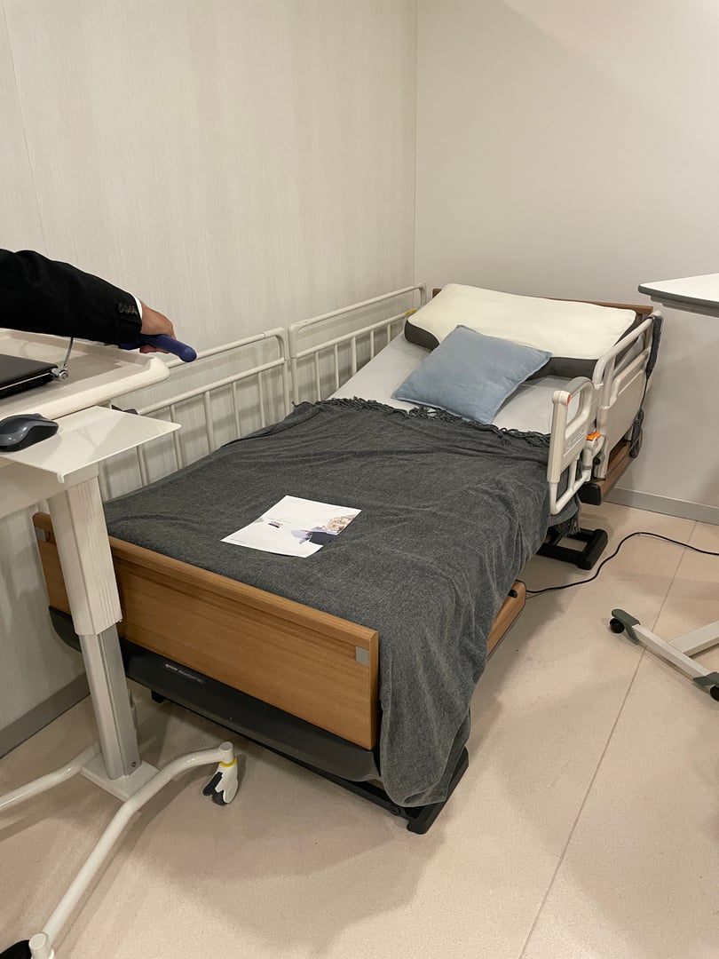 Hospital bed with gray blanket, pillow, and a small bedside table, located in a clean, modern room setting.