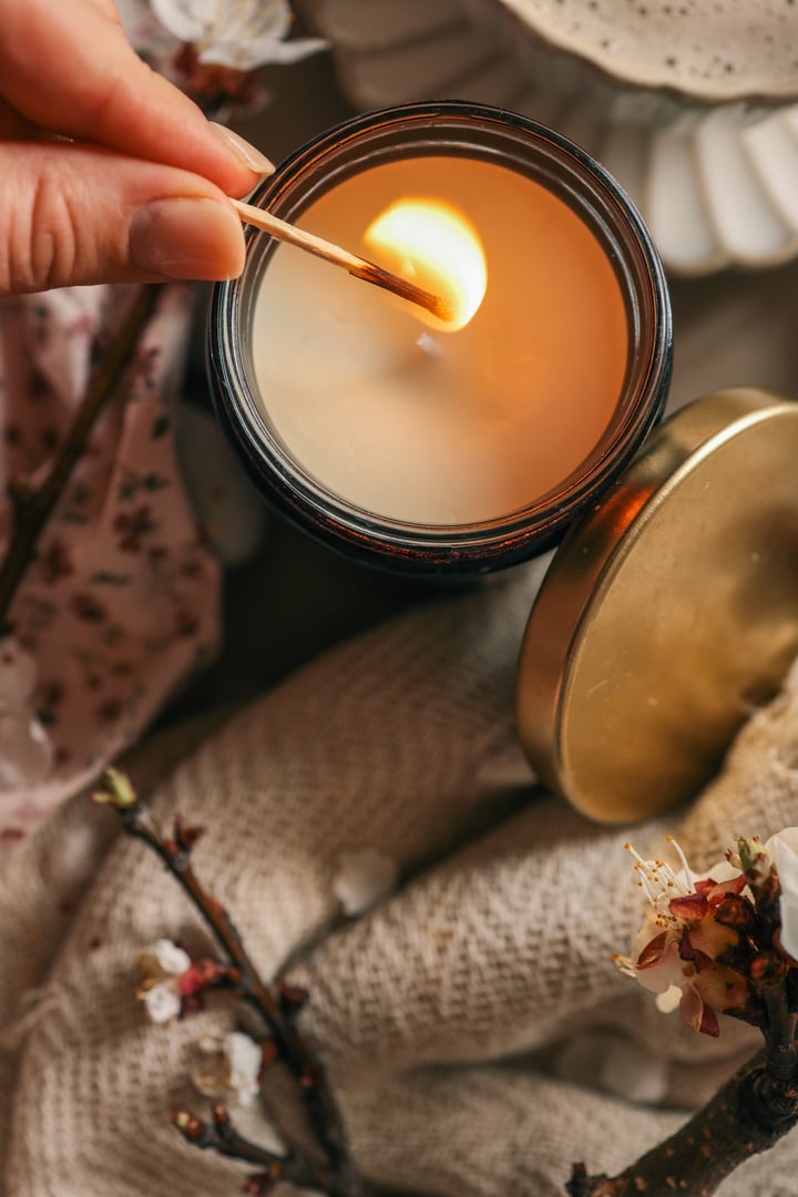 Lighting a soy candle in a dark glass jar with a matchstick. Cozy ambiance with soft knitted fabric, cherry blossoms, and delicate spring mood.