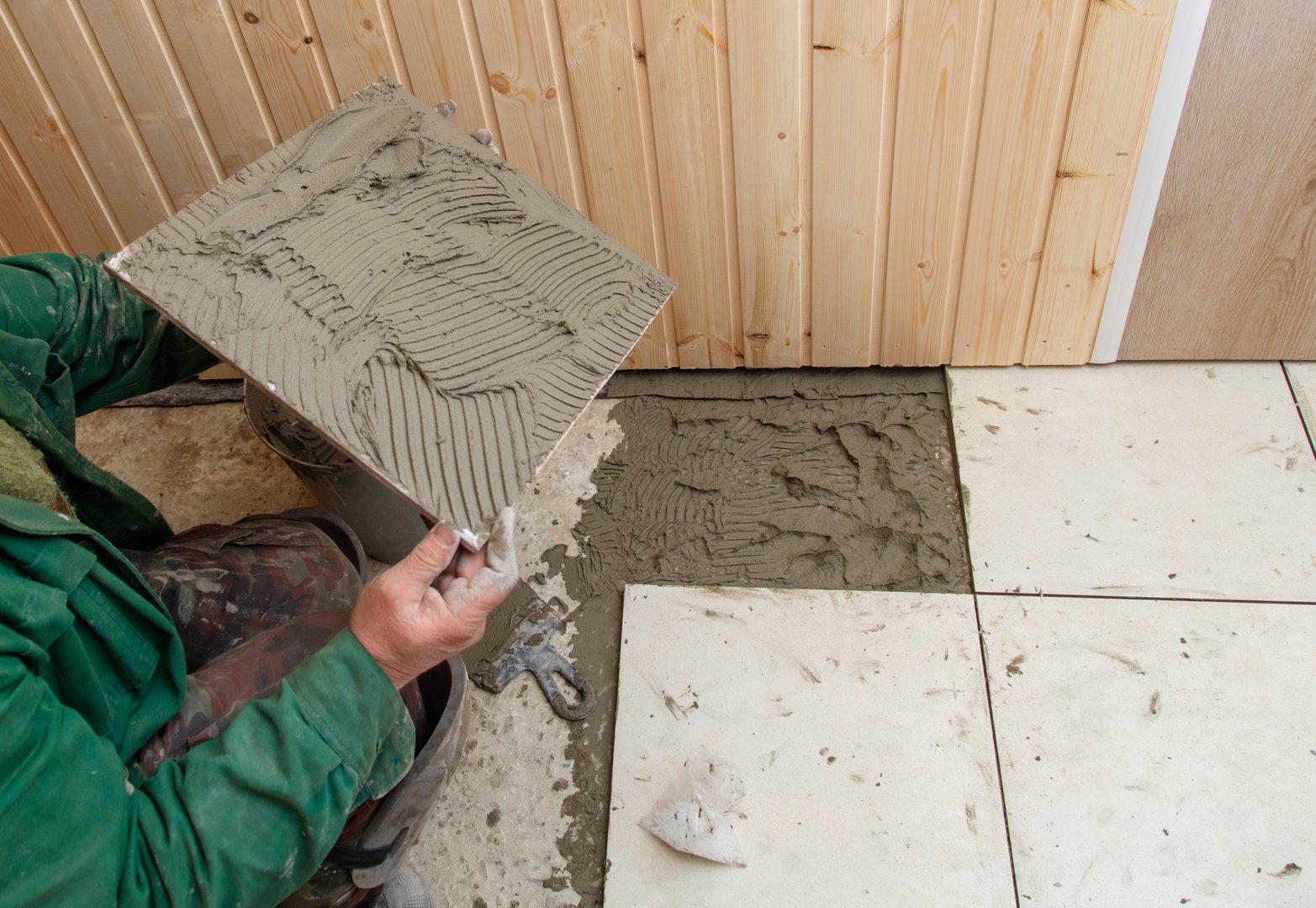 A man is laying tiles on a floor. He is wearing a green shirt and is holding a trowel