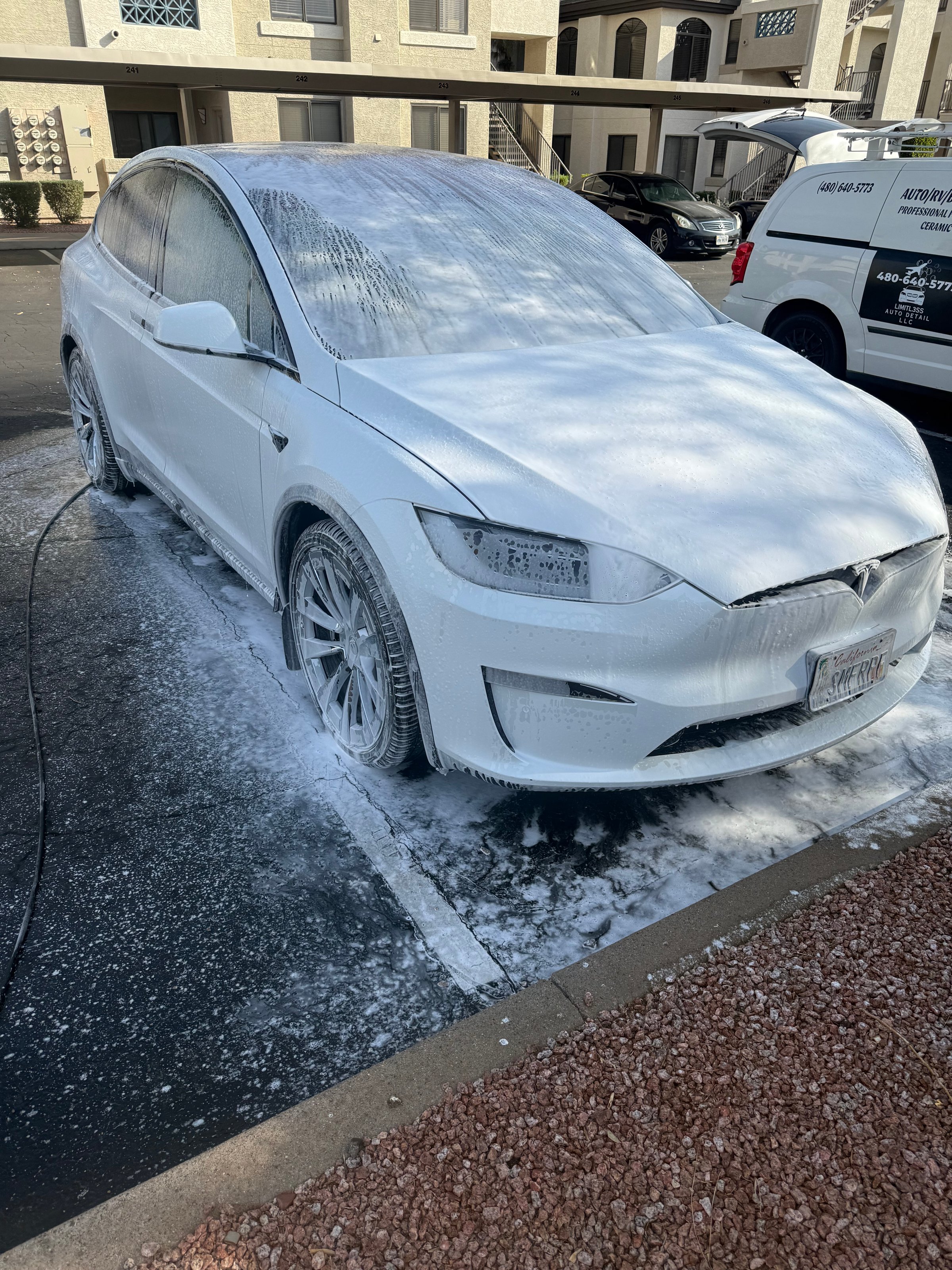 White electric car covered in white foam after a wash, parked in a lot near residential buildings.