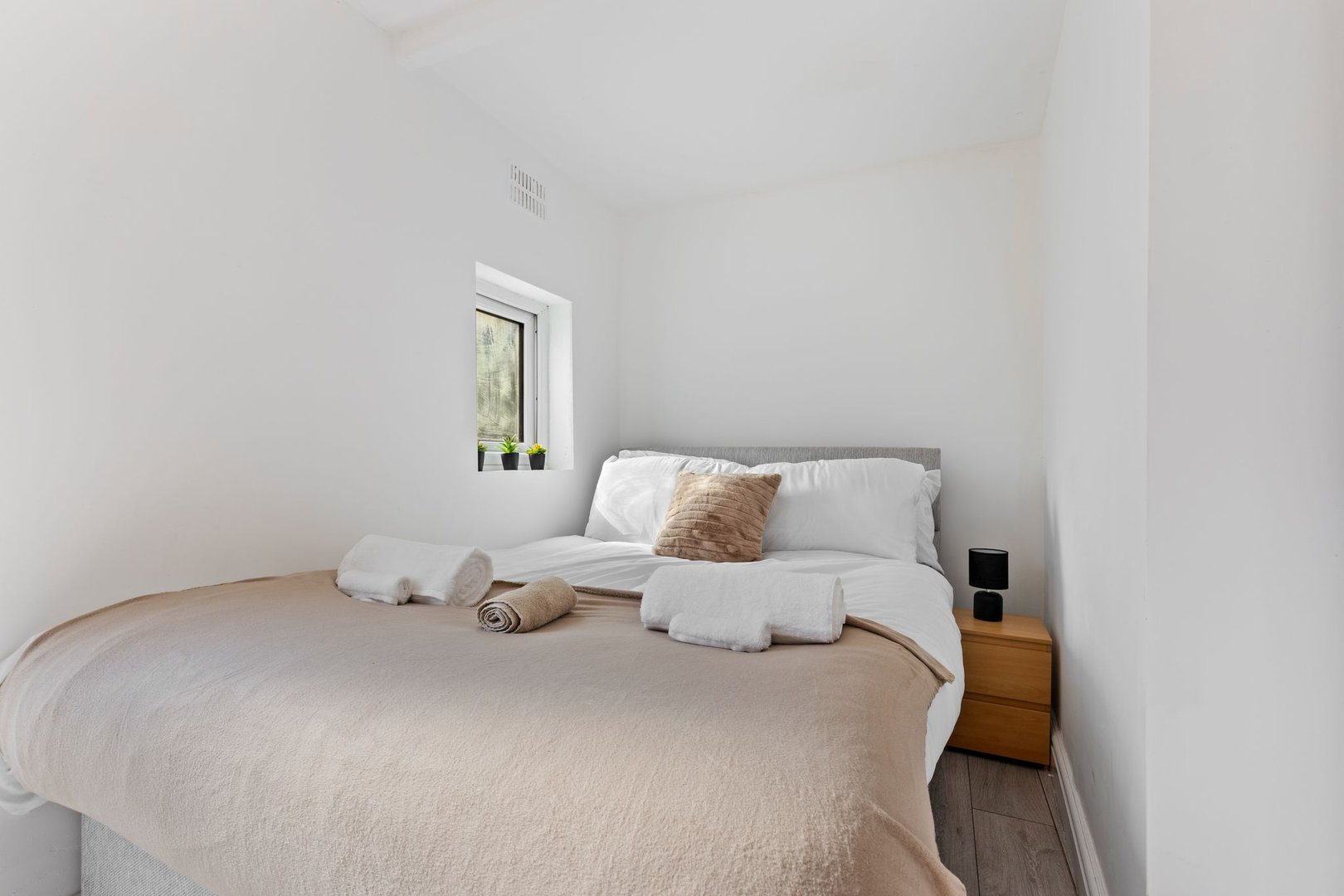 Minimalist bedroom with double bed, beige bedding, towels, small window, and potted plant on windowsill.