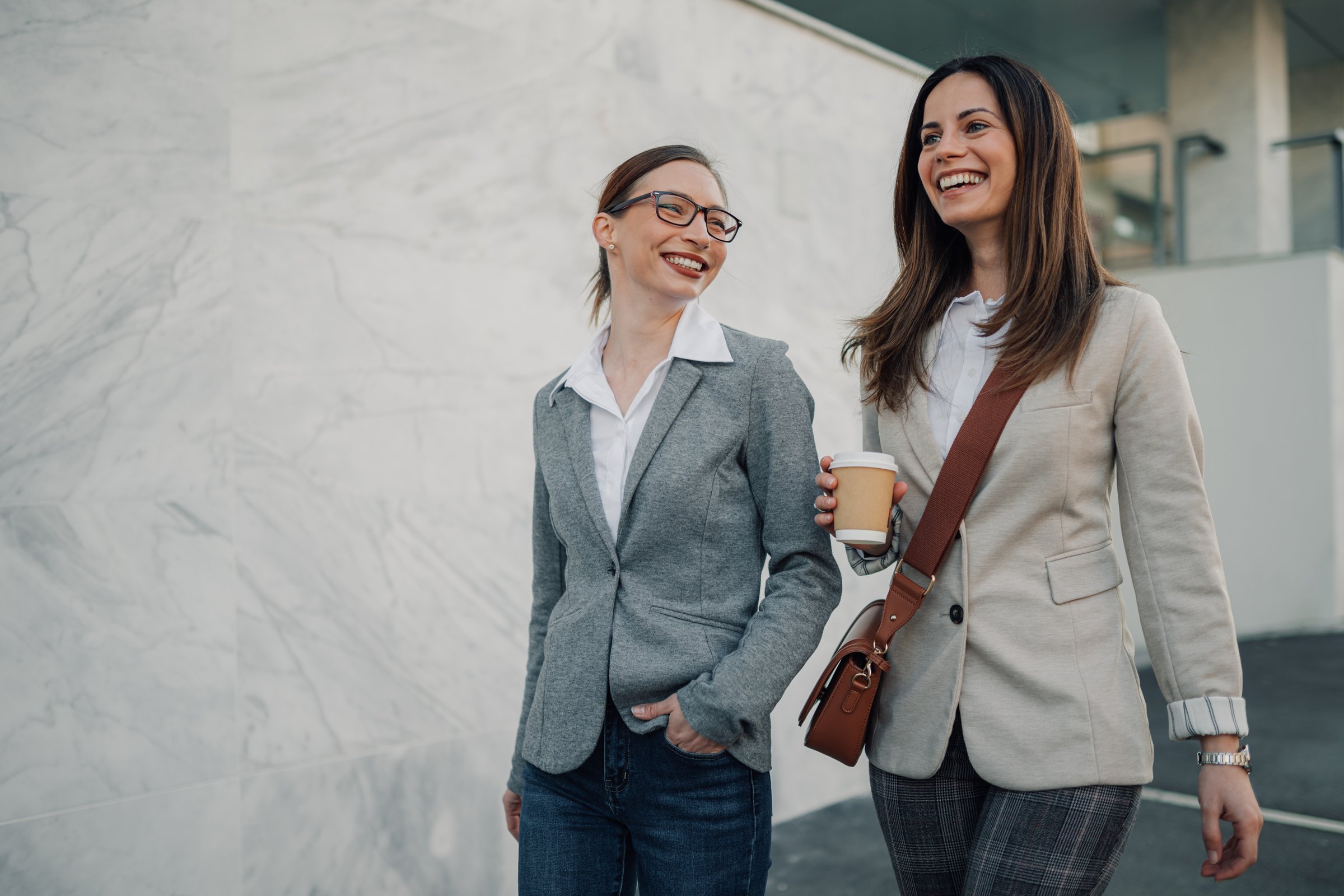 Two businesswomen are walking and talking on a city street, enjoying a coffee break together