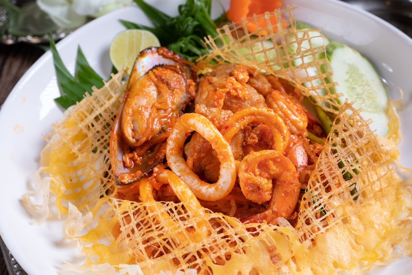 Seafood pasta topped with a crispy lattice garnish, served with lime, cucumber slices, and herbs on a white plate.