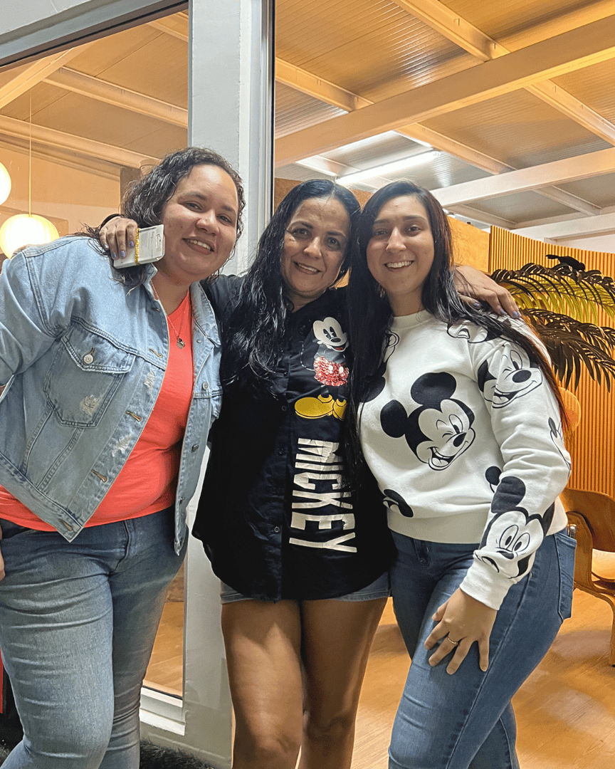 Three women smiling, with two wearing Mickey Mouse-themed clothing, standing together indoors near a window.