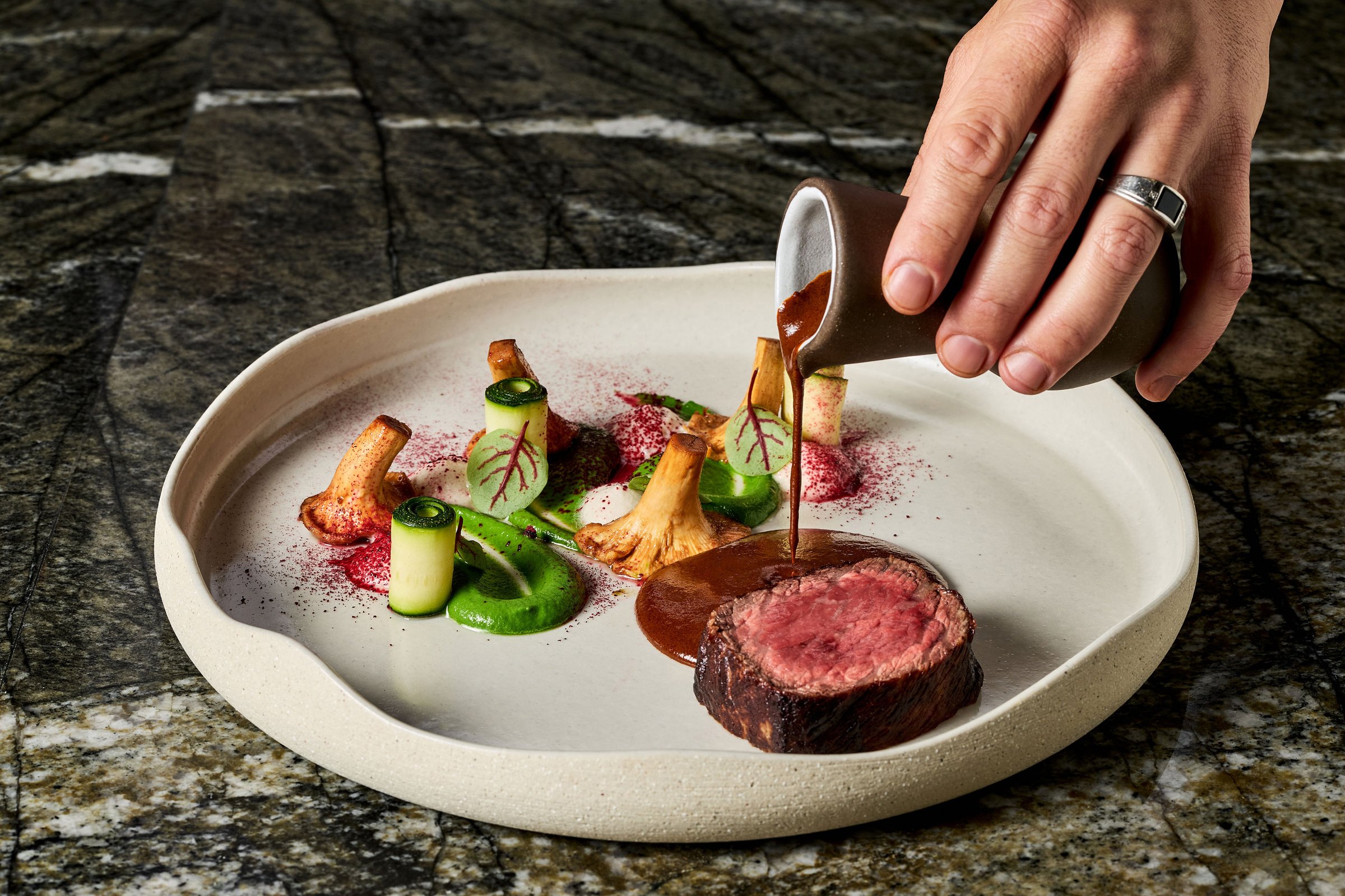 Professional chef hand pouring sauce over medium-rare beef filet with mushrooms and vegetables. Fine dining culinary art presentation on ceramic plate.