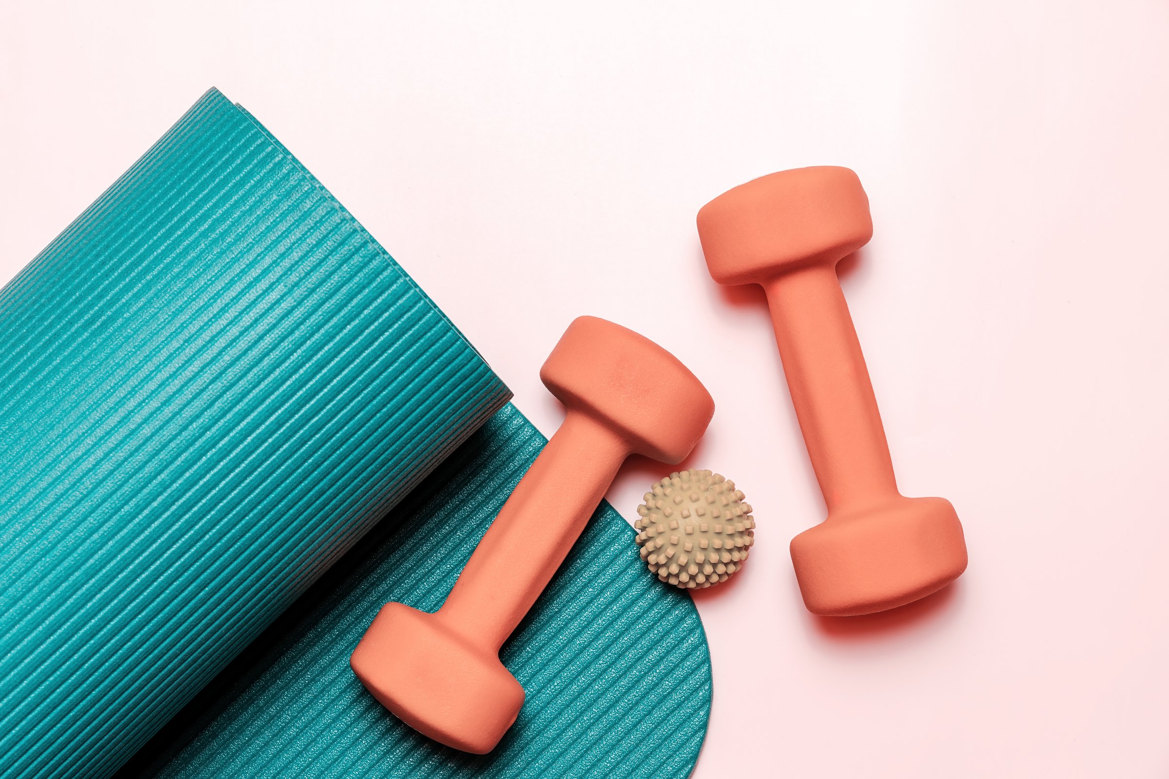 Teal yoga mat is partially rolled up, next to two coral dumbbells and a beige massage ball. Perfect for health and fitness concepts