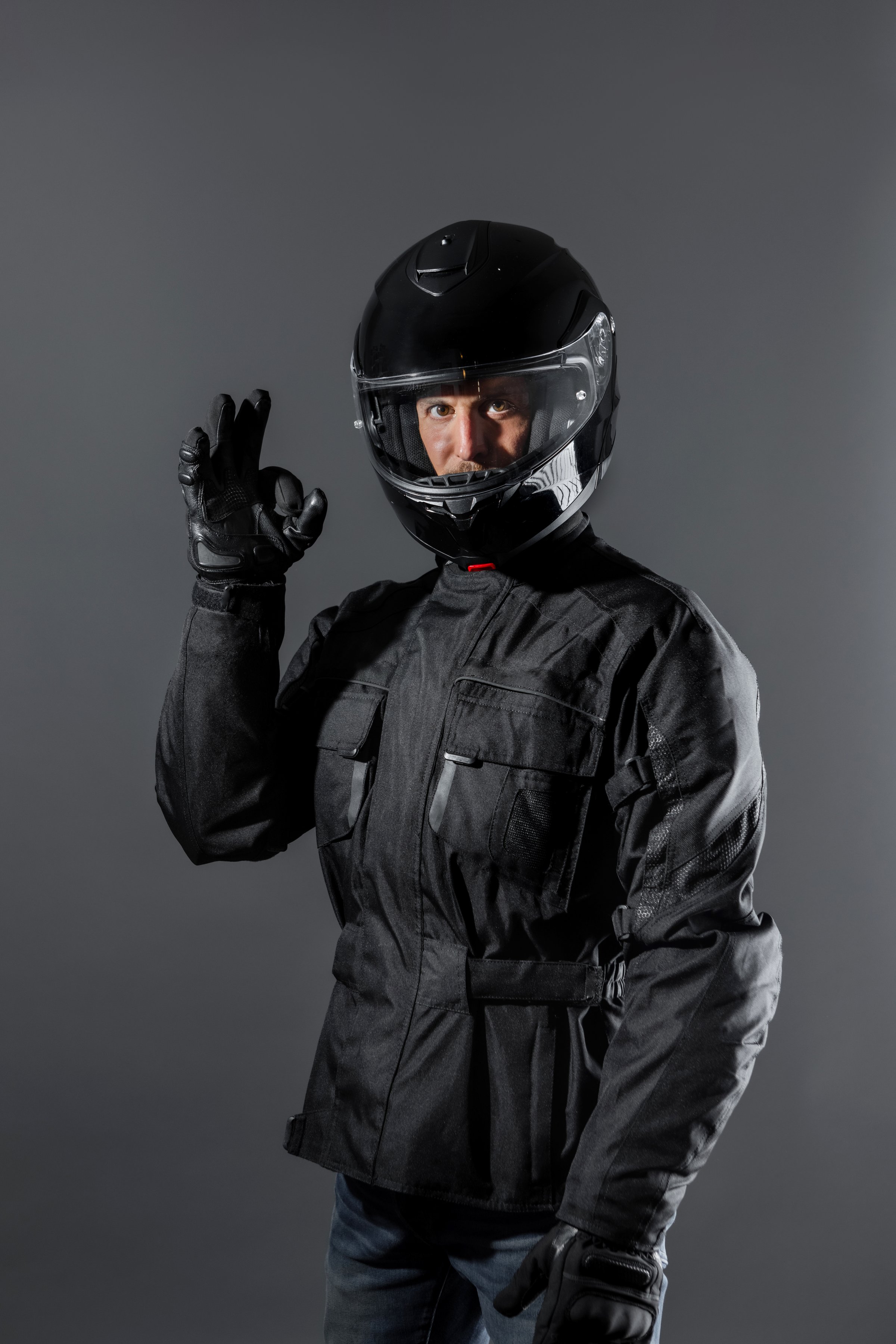 Safe ride concept. Confident Man wearing protective motor black clothing doing ok sign in studio. Motorcycling equipment. Studio shot, black background, indoors.