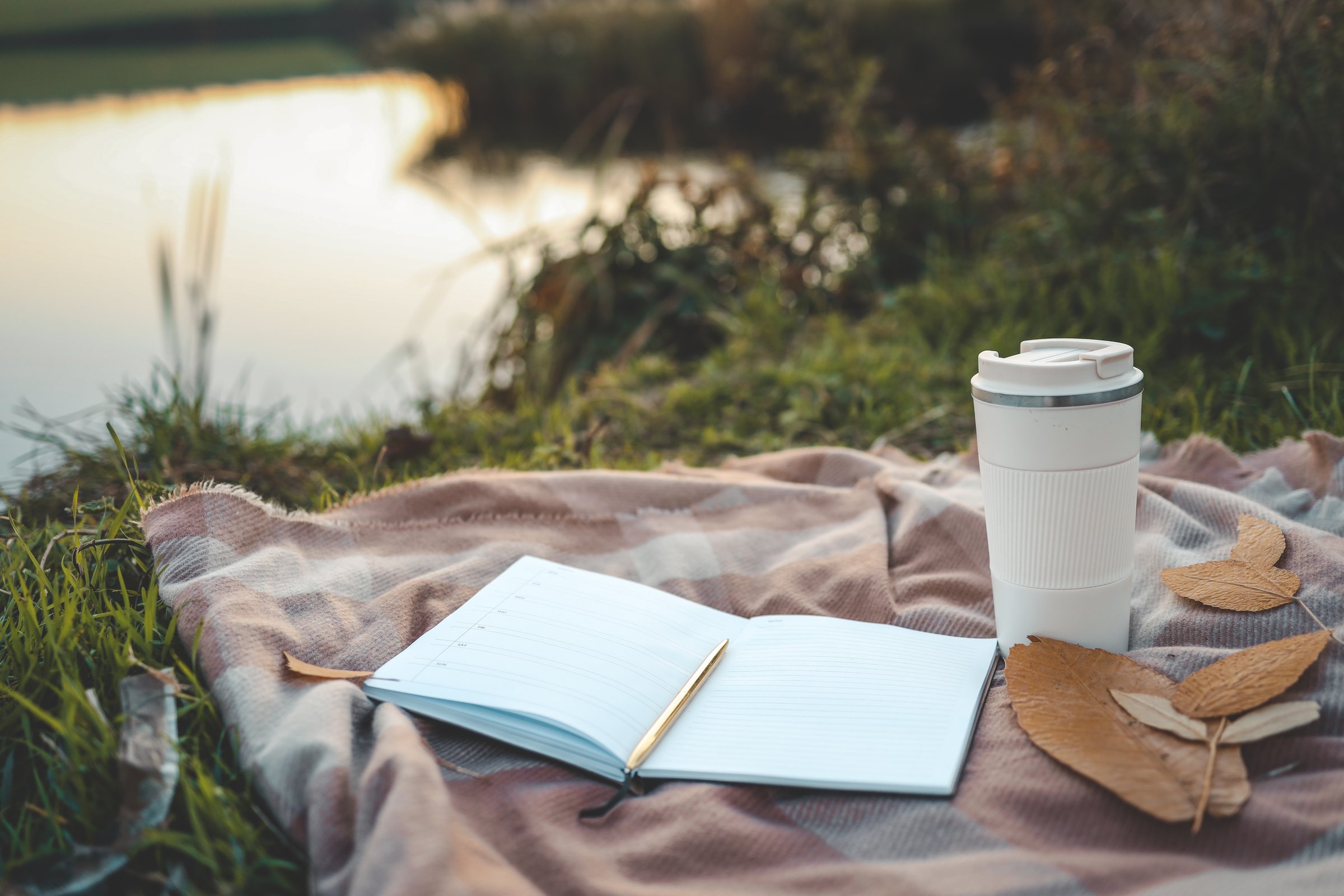 Open planner and reusable coffee cup on a plaid blanket near a peaceful lake at sunset