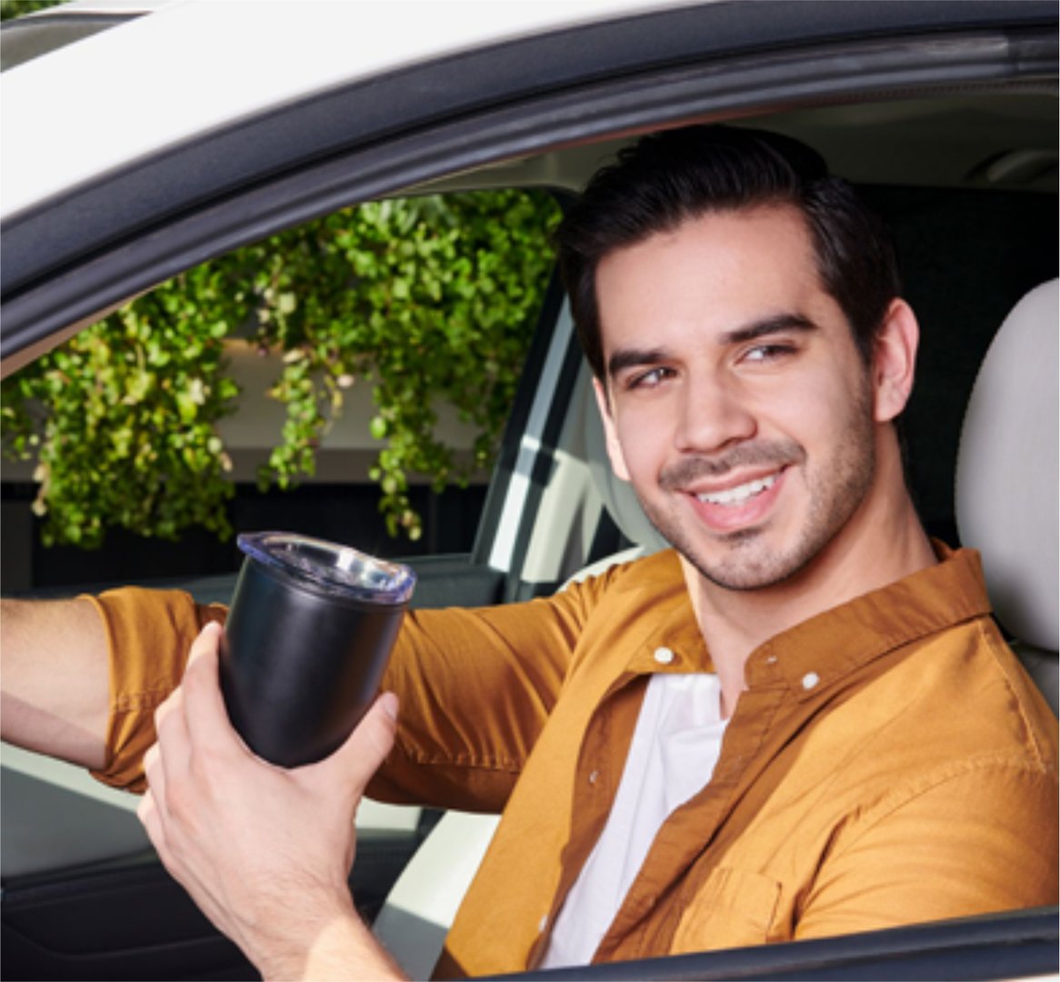 Man smiling in car holding a black travel mug, wearing a brown jacket. Greenery visible outside the window.