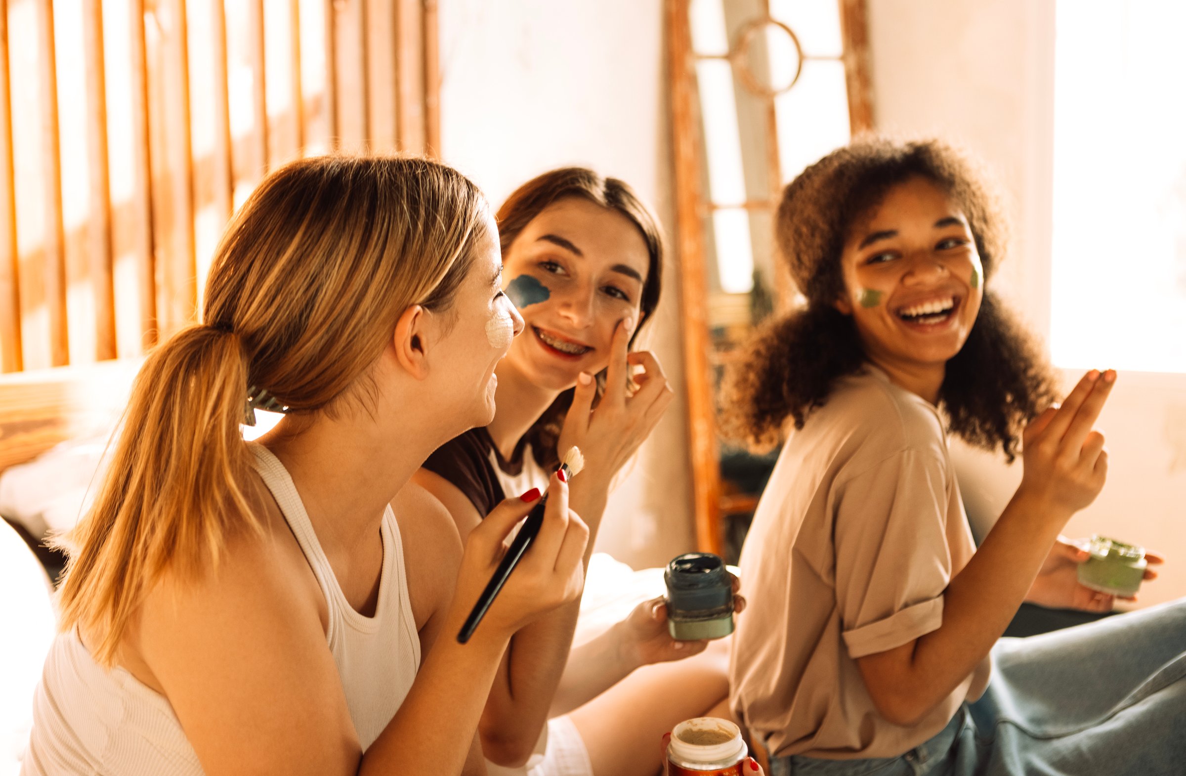 Three charming teenagers of different races apply cosmetic masks to their faces in the bedroom. Female multiethnic friends have fun together at home. Cute funny college girls take care of their skin.