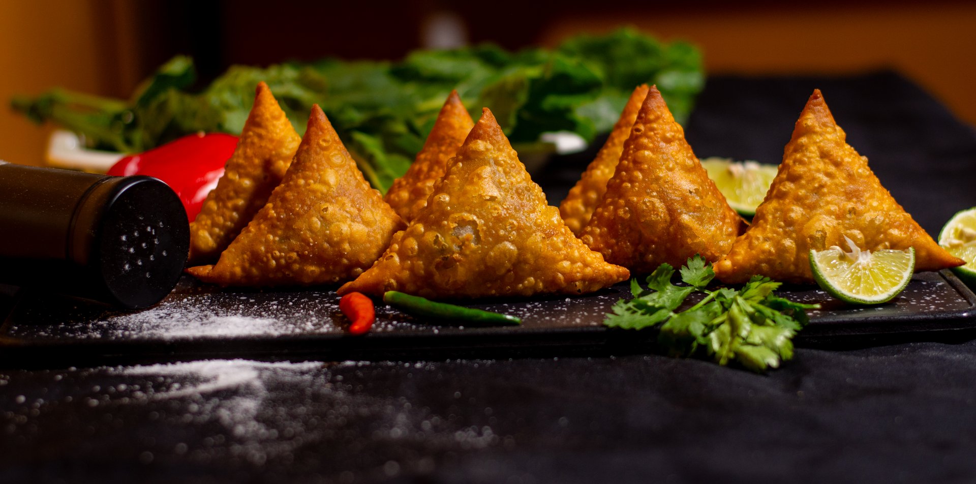 Four samosas on a black tray with lime slices, red and green chilies, and cilantro, set against a dark background.