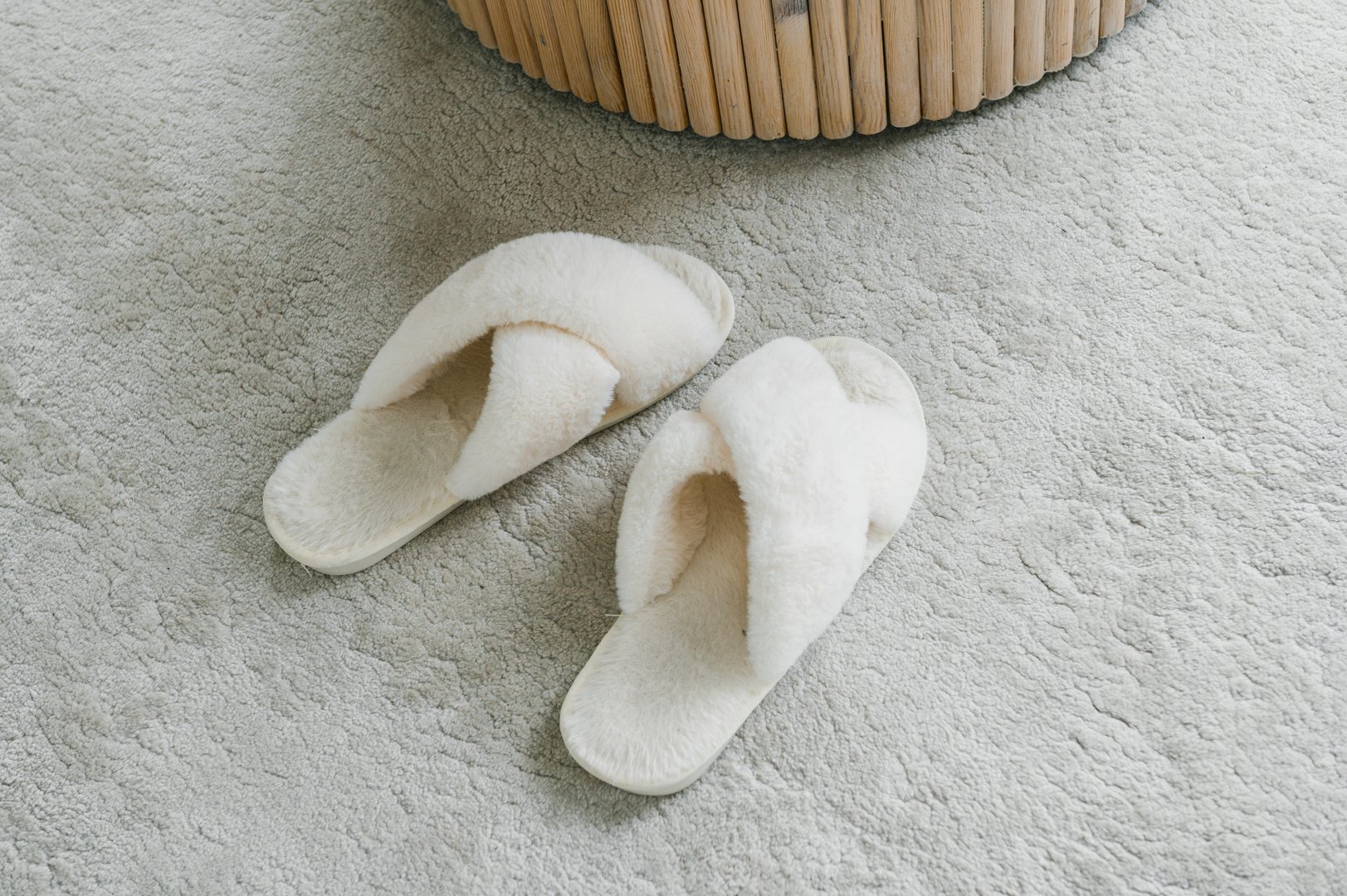 A pair of soft white slippers rests beside a stylish wicker basket, placed neatly on a textured plush carpet, evoking a sense of home comfort